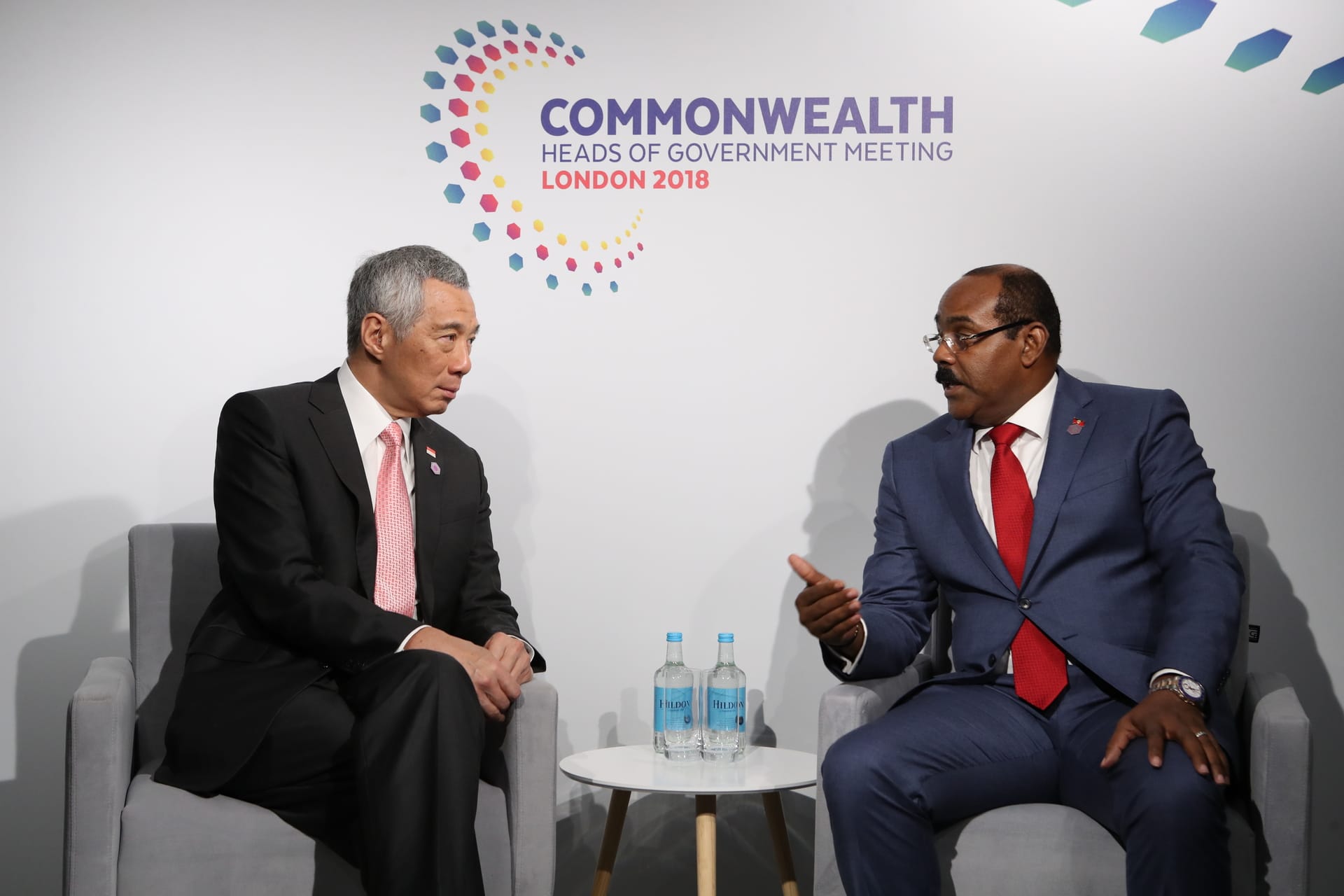 Two men in suits seated, talking; "Commonwealth Heads of Government Meeting London 2018" backdrop.