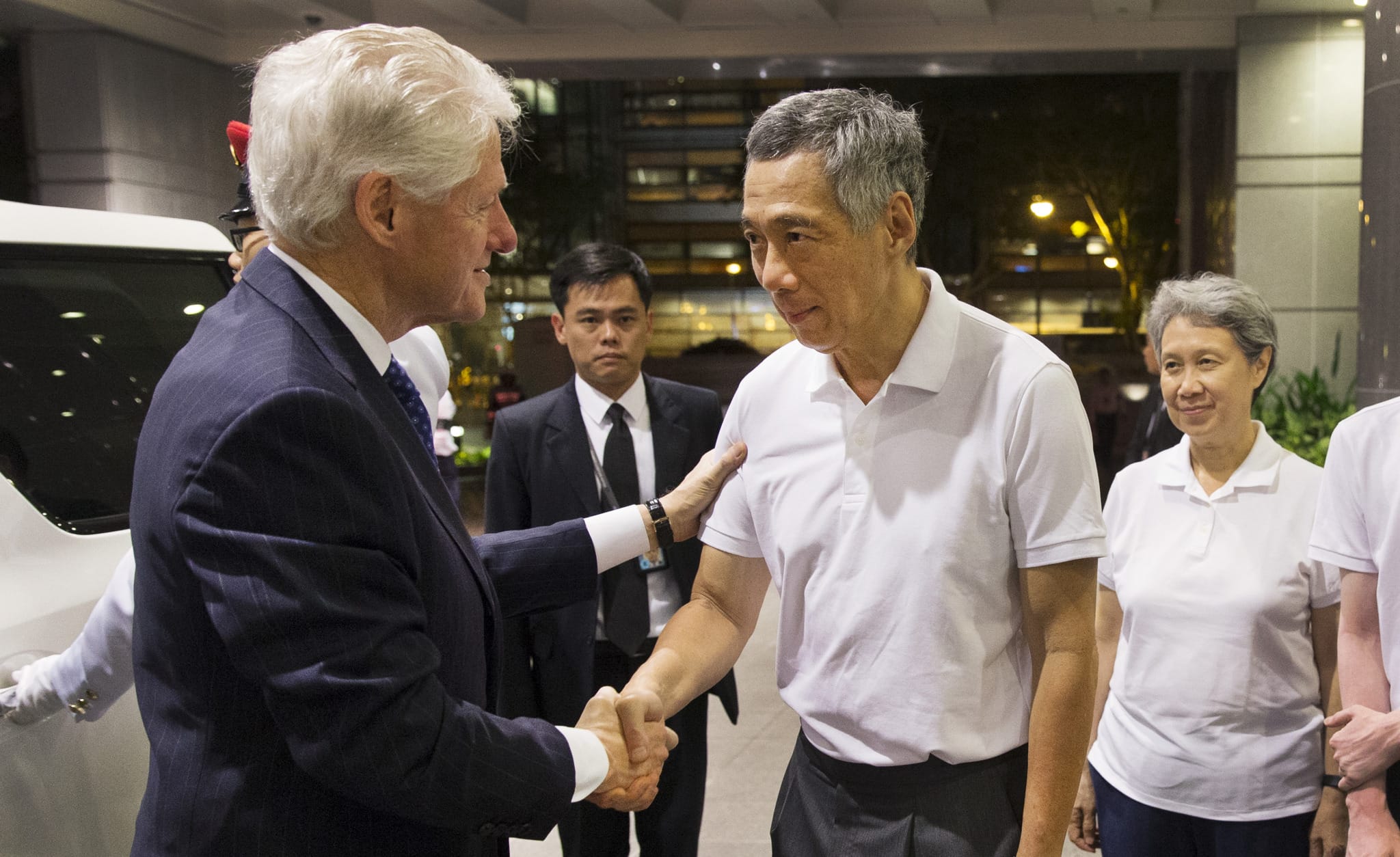 Bill Clinton shakes hands with Lee Hsien Loong, surrounded by others in suits and polo shirts.