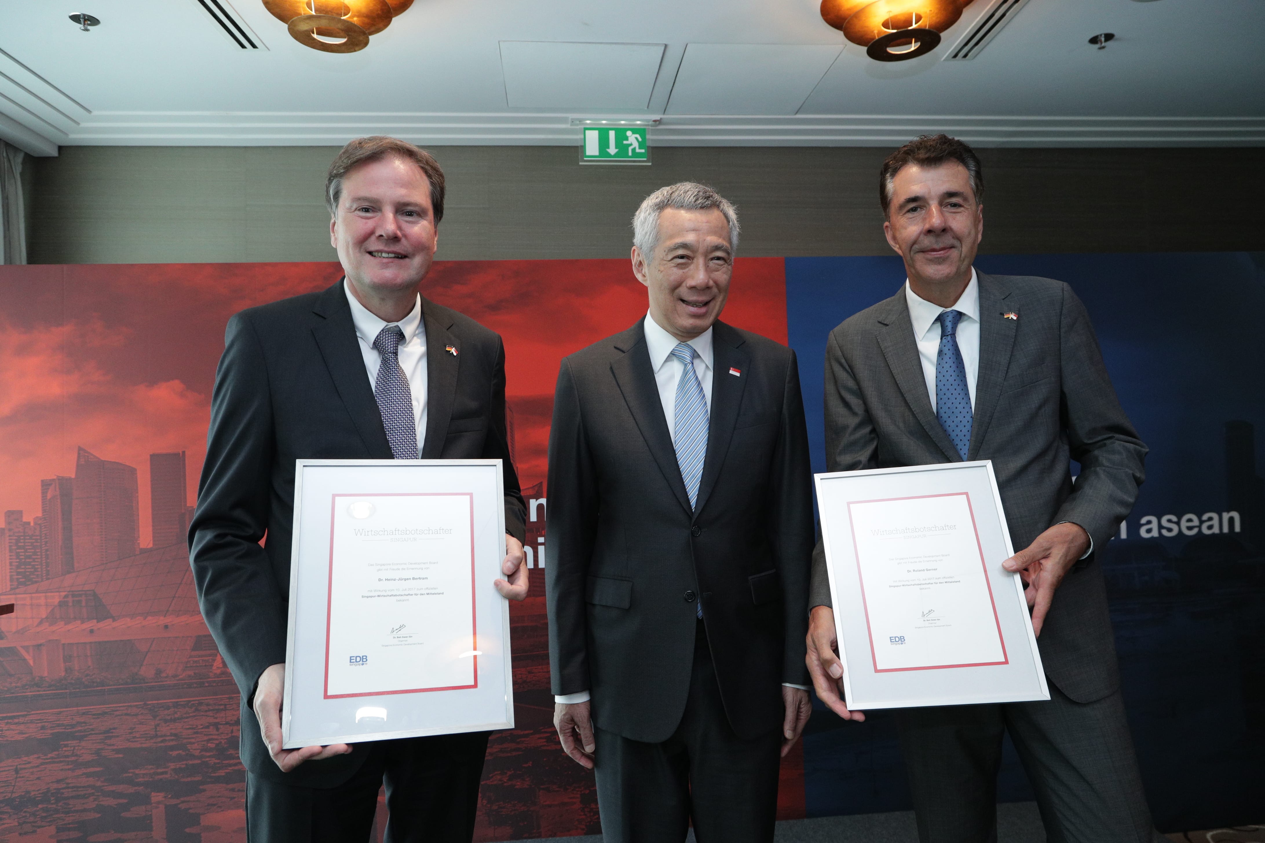 Three men in suits holding framed certificates in front of a red/blue background. Lee Hsien Loong is in the middle.