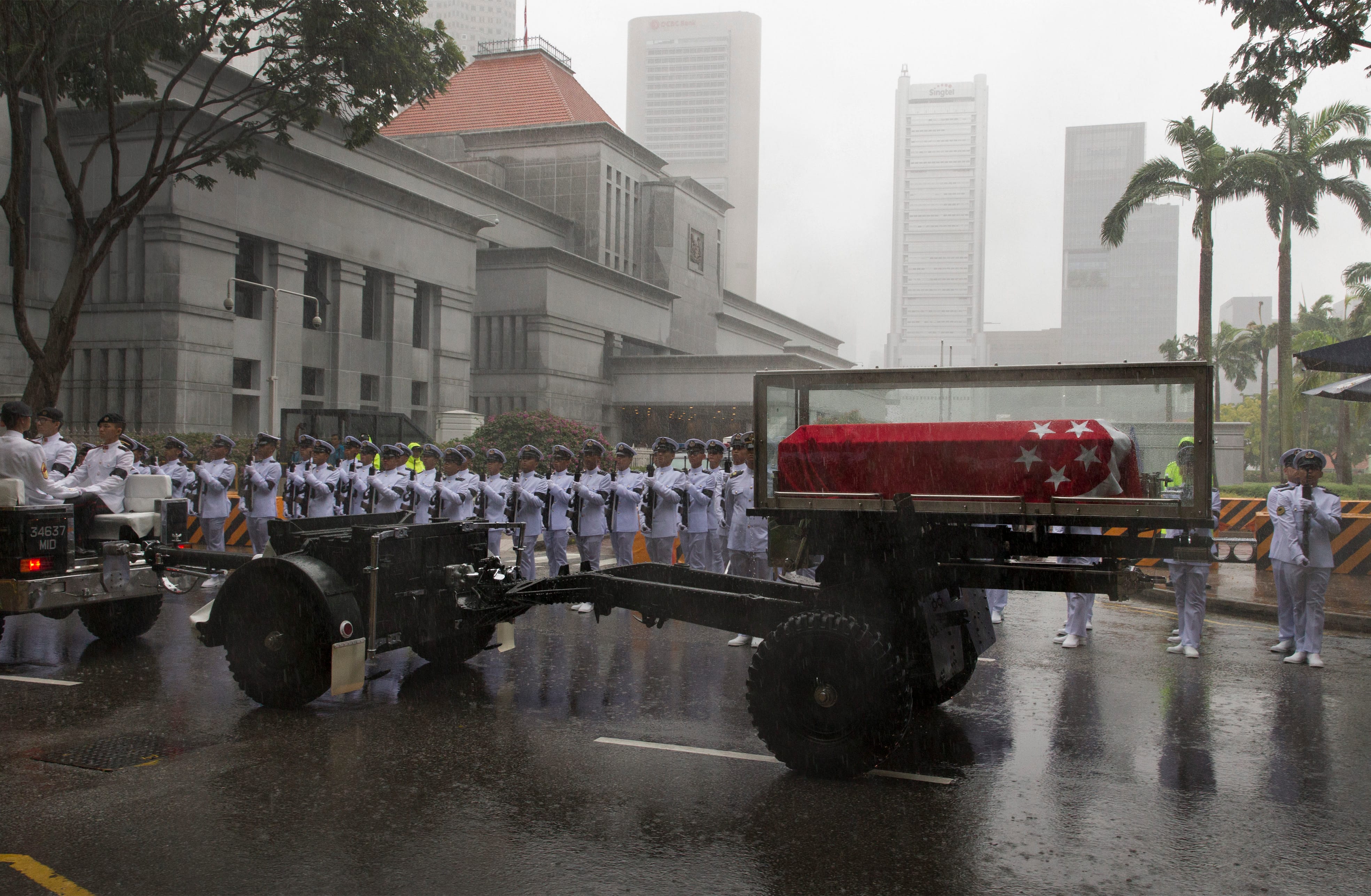 Raining scene shows Singapore flag-draped coffin on carriage towed by military personnel in white uniforms.