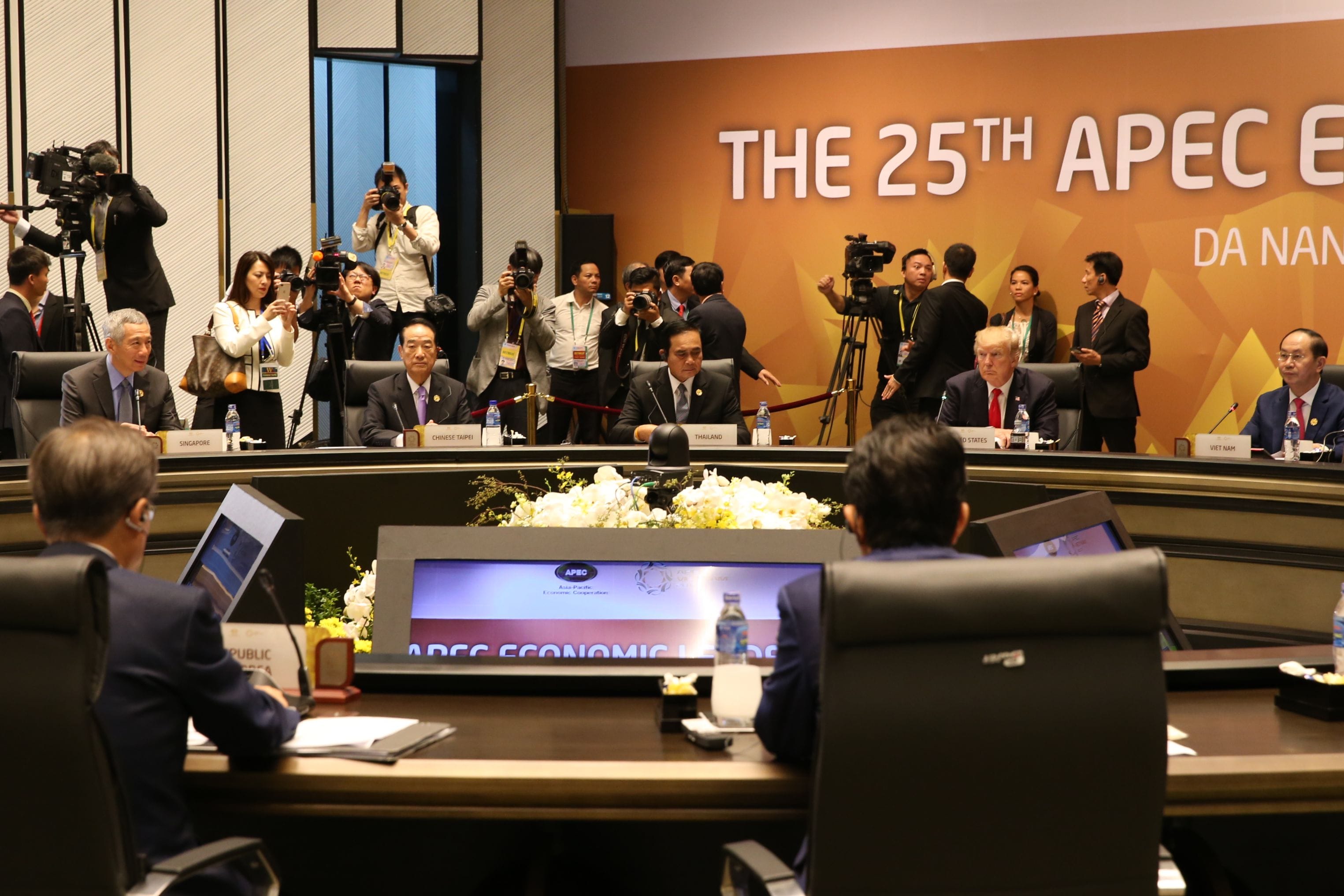 APEC Economic Leaders' Meeting with global leaders, cameras flashing and orange backdrop.