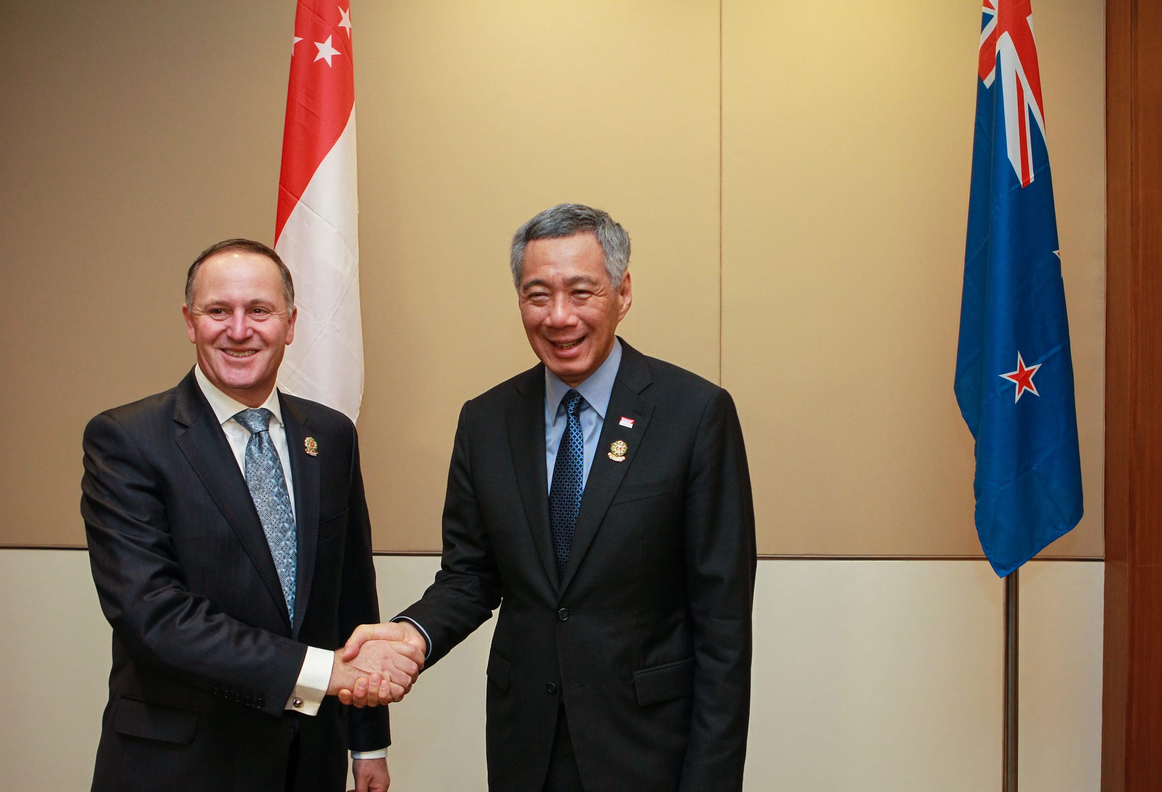 Two men in suits shaking hands, with Singapore and New Zealand flags in the background.