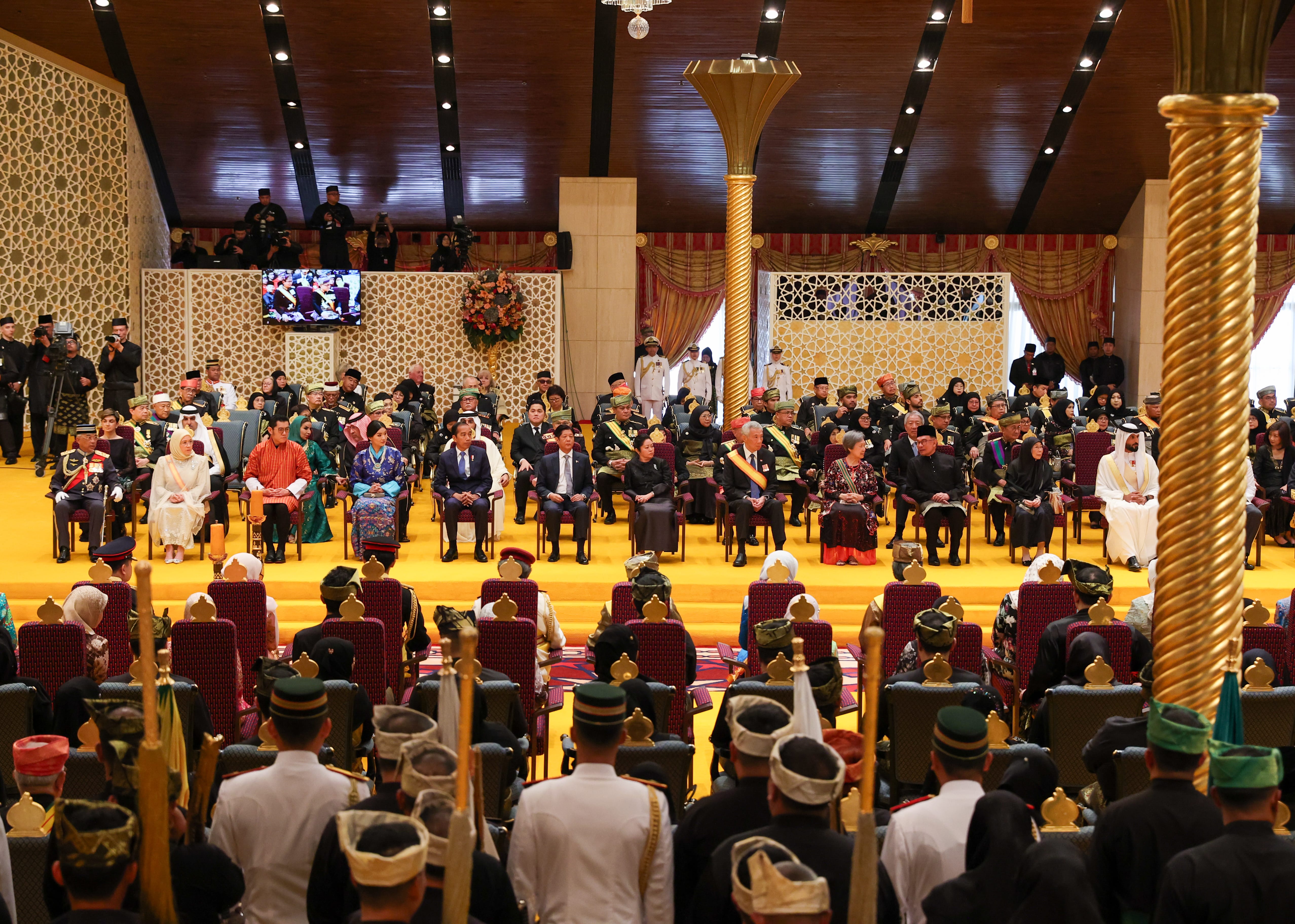 Large audience seated in rows beneath an ornate, lit ceiling and geometric wall patterns.