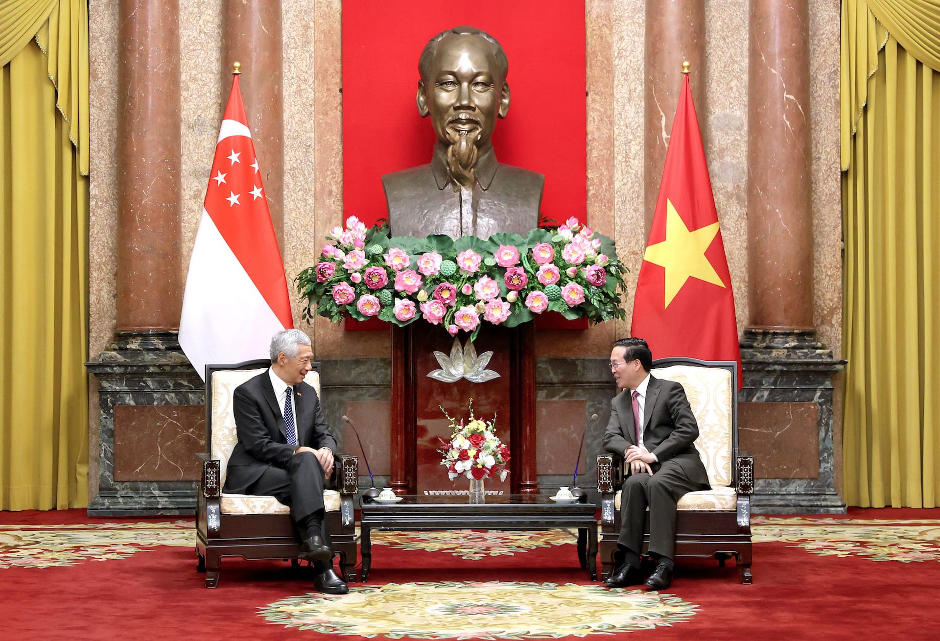 Two men in suits sit facing each other between Singapore & Vietnam flags and Ho Chi Minh bust.