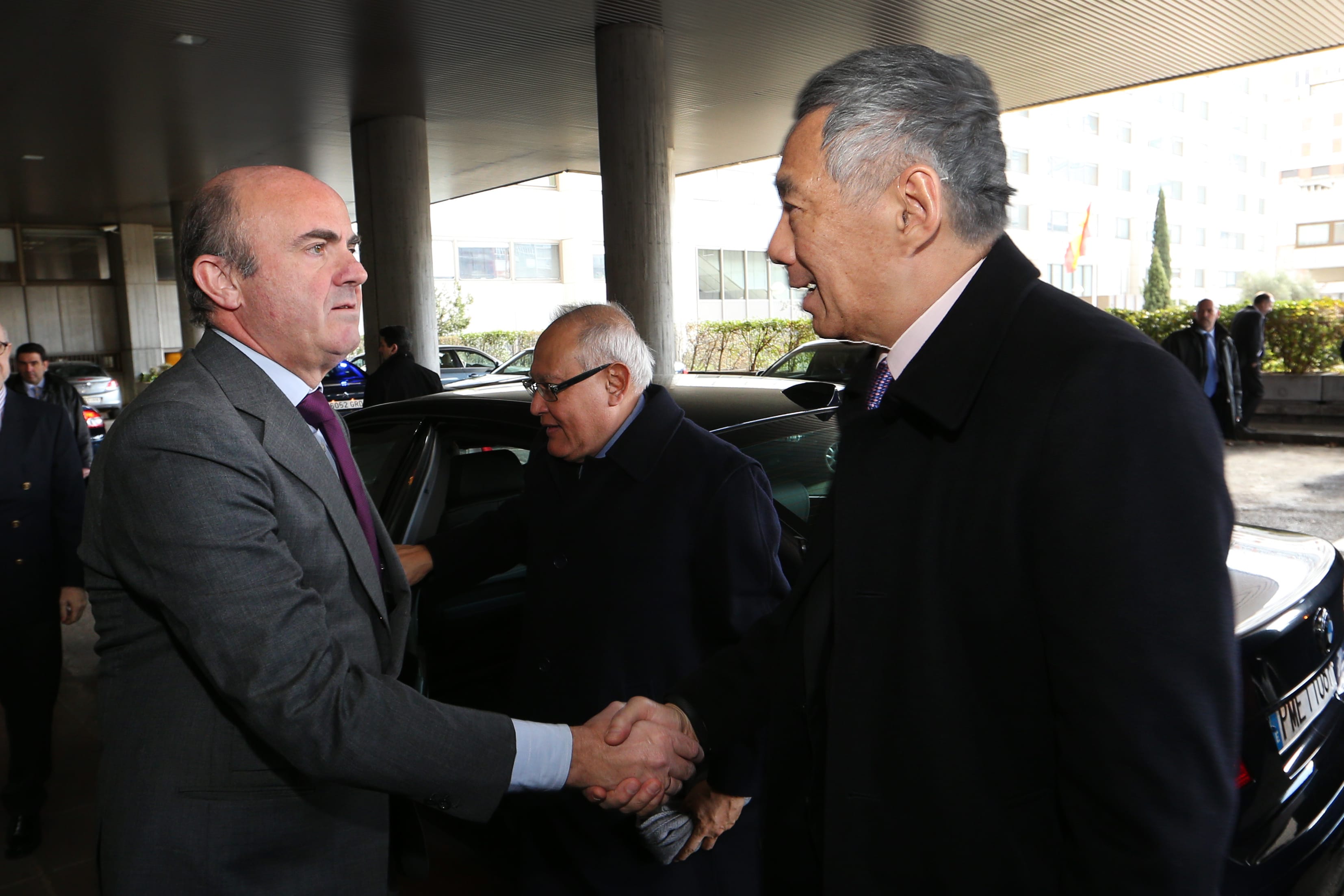 Two men in suits shaking hands near a black car under a building's overhang.