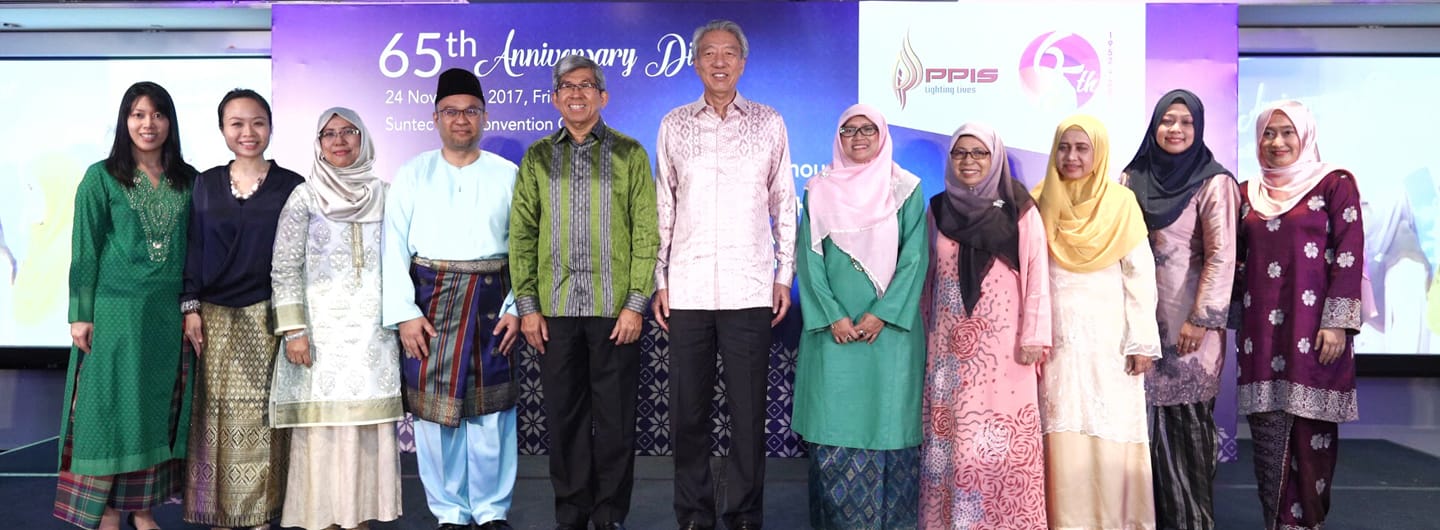 People standing in a row against a 65th Anniversary backdrop. Varied colors and patterns in their clothing.