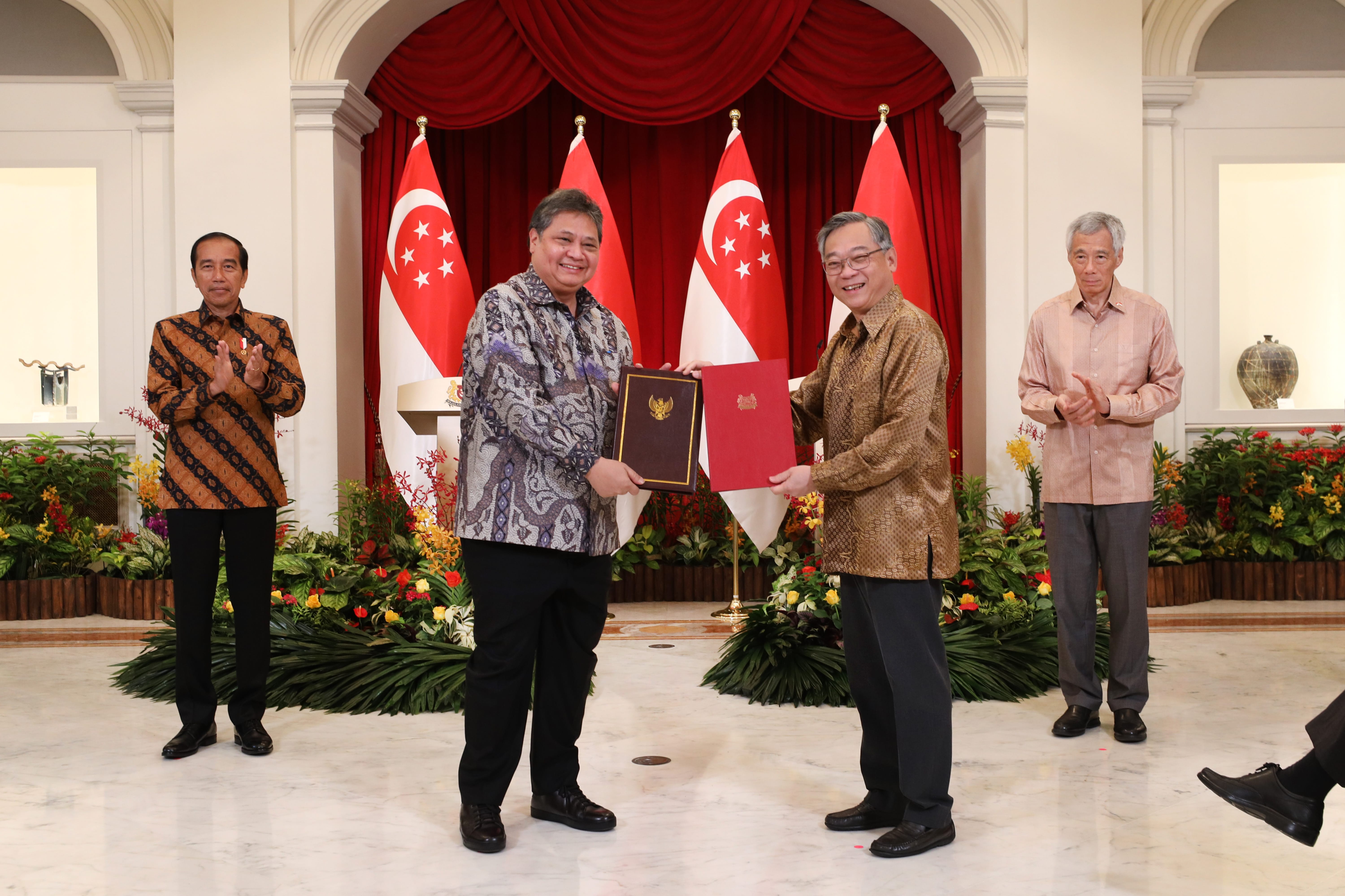 Two men exchanging documents in front of Indonesian and Singaporean flags. Other men are standing by.