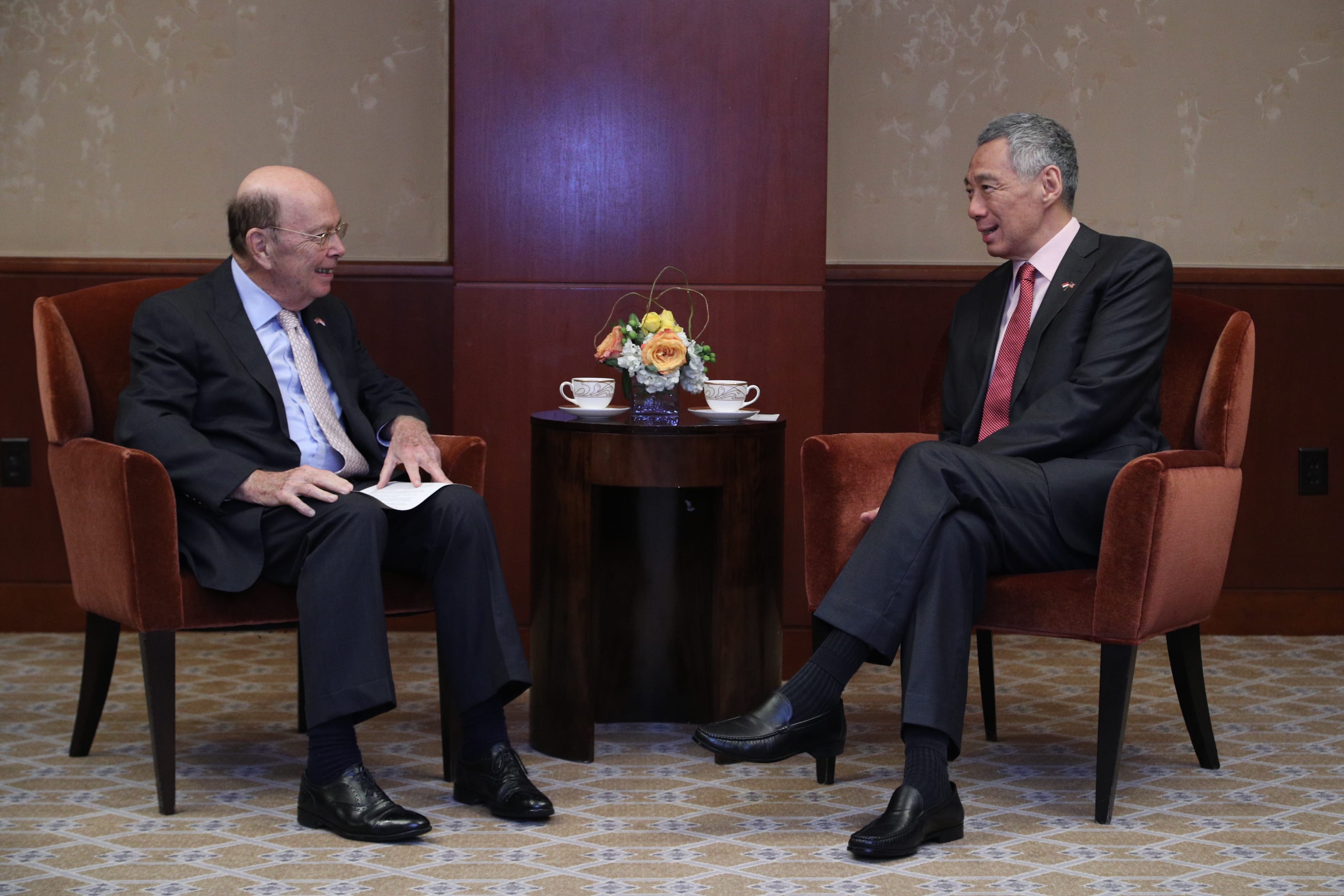Wilbur Ross and Lee Hsien Loong converse, seated in armchairs across from a table with flowers and tea.