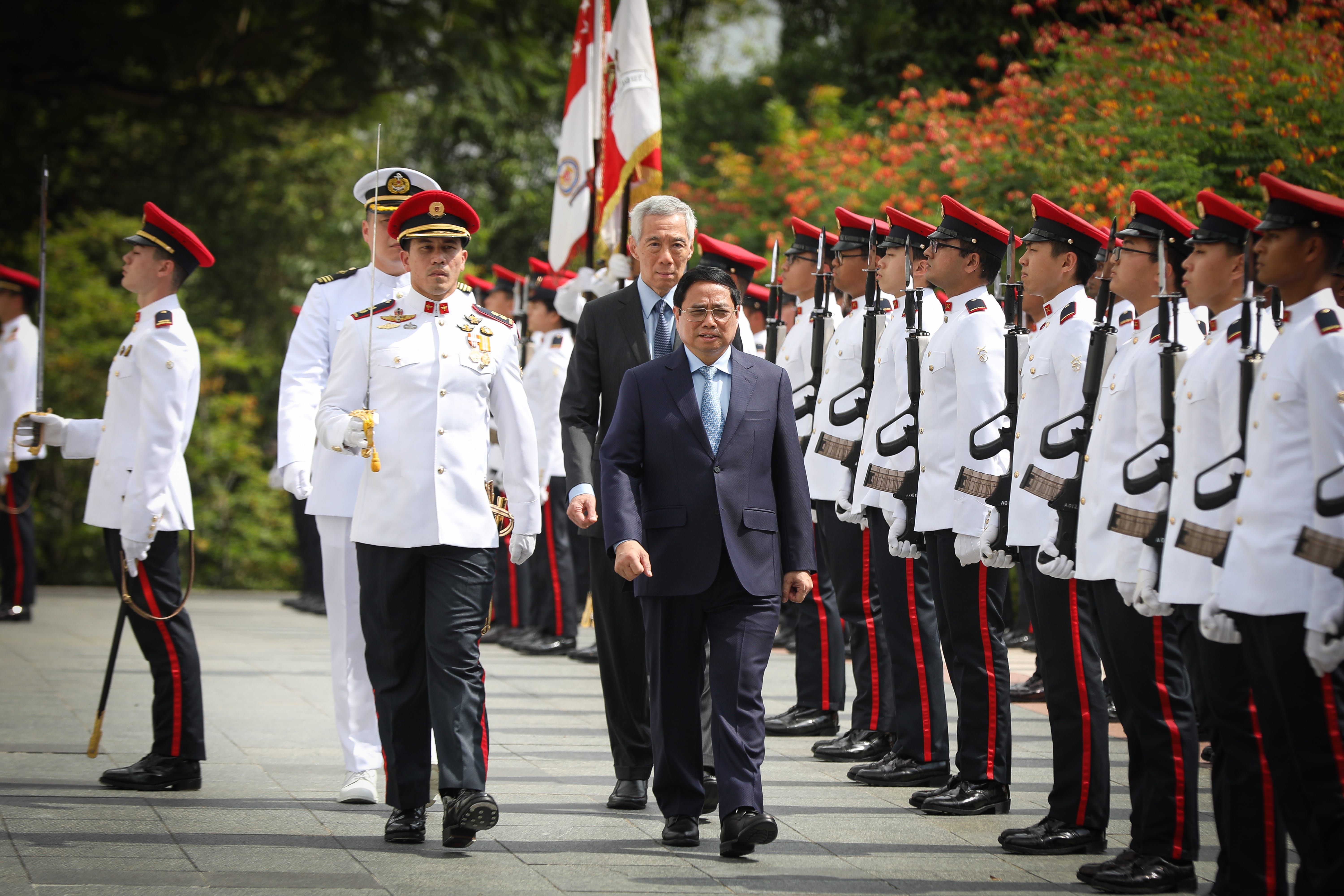 Men in suits walk past a military honor guard in white uniforms with red trim. Flags are visible.