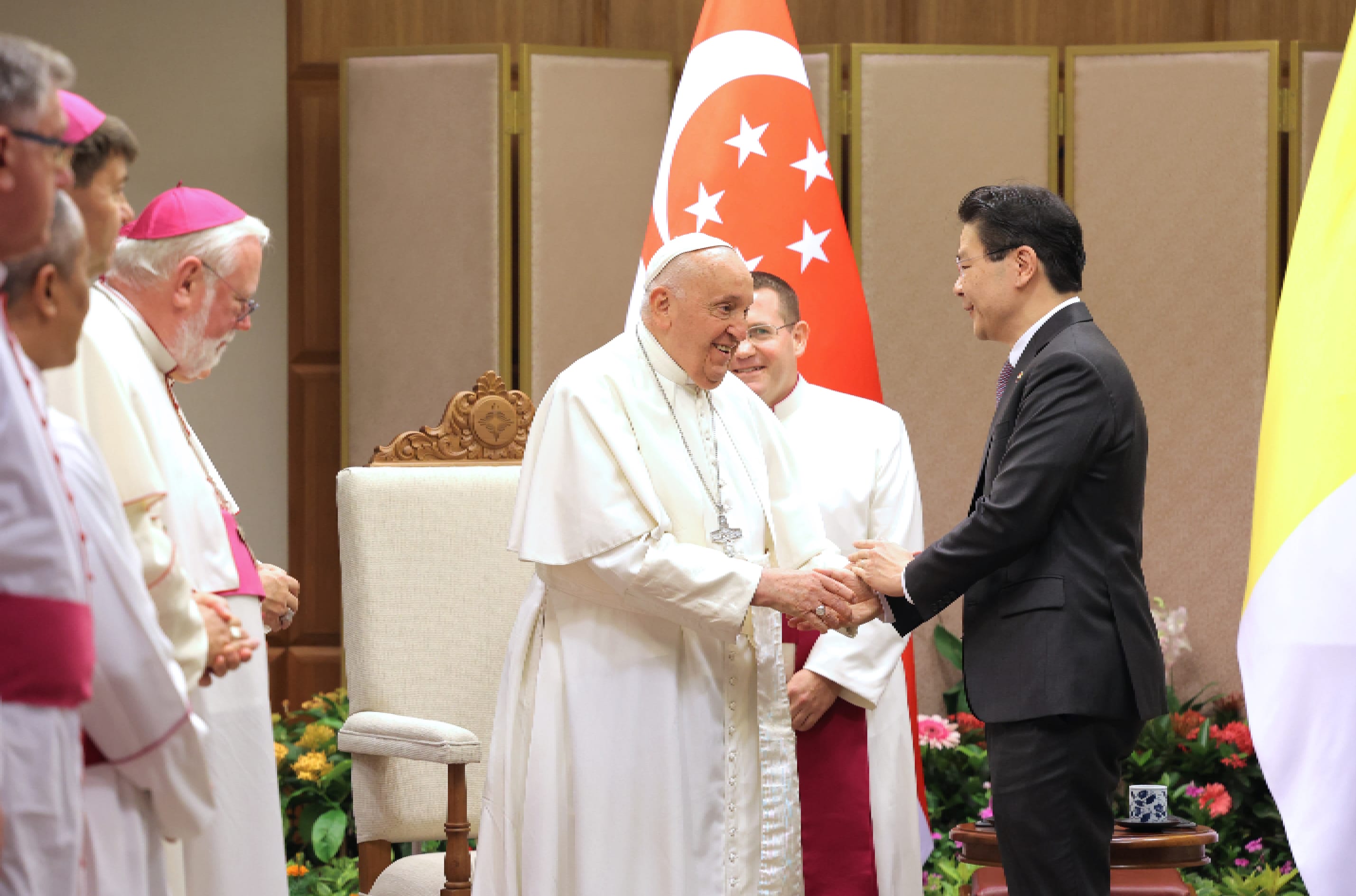 Pope Francis in white robes shaking hands with a man in a black suit. Singapore flag visible.