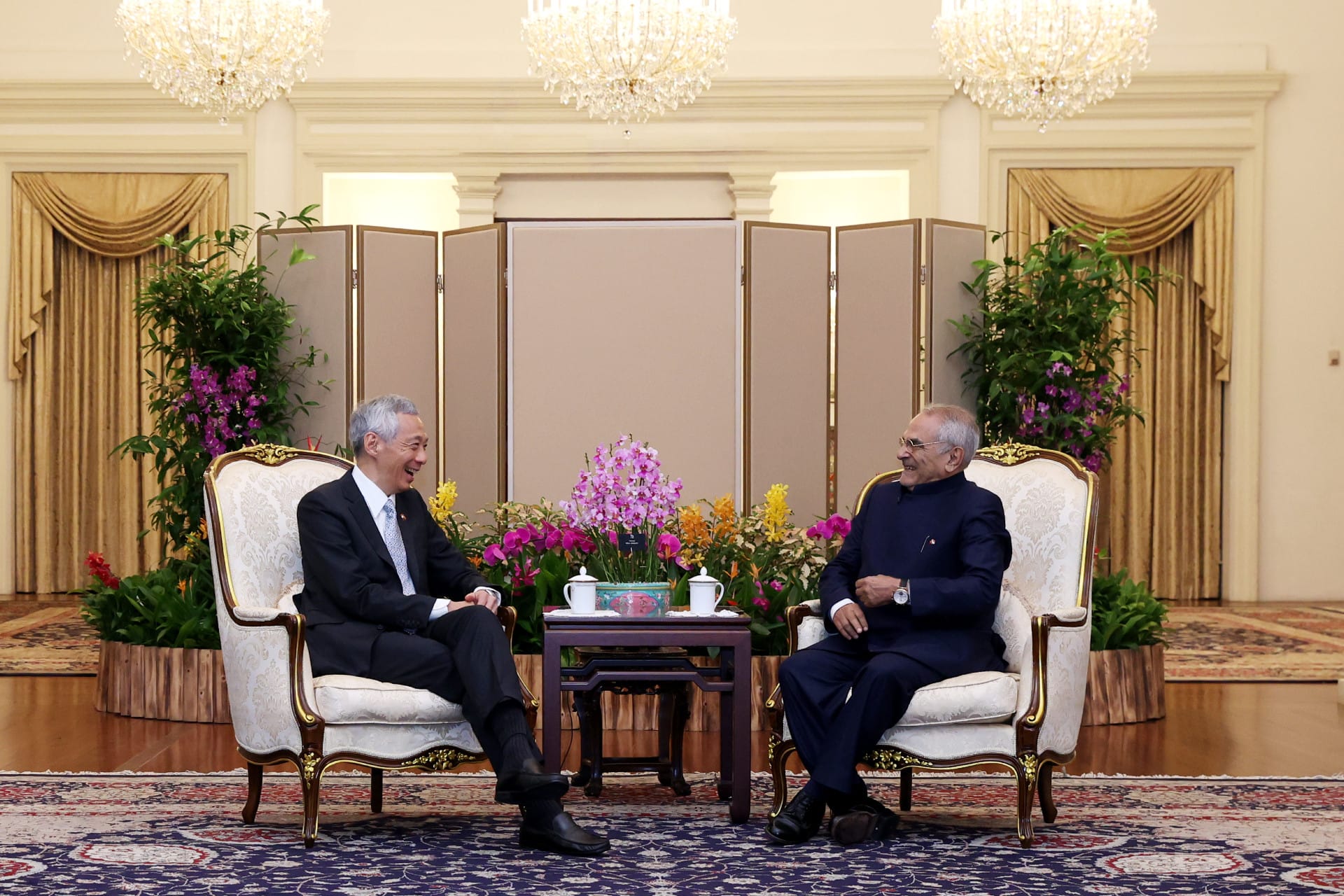 Two men in suits seated in ornate chairs facing each other across a table with flowers.