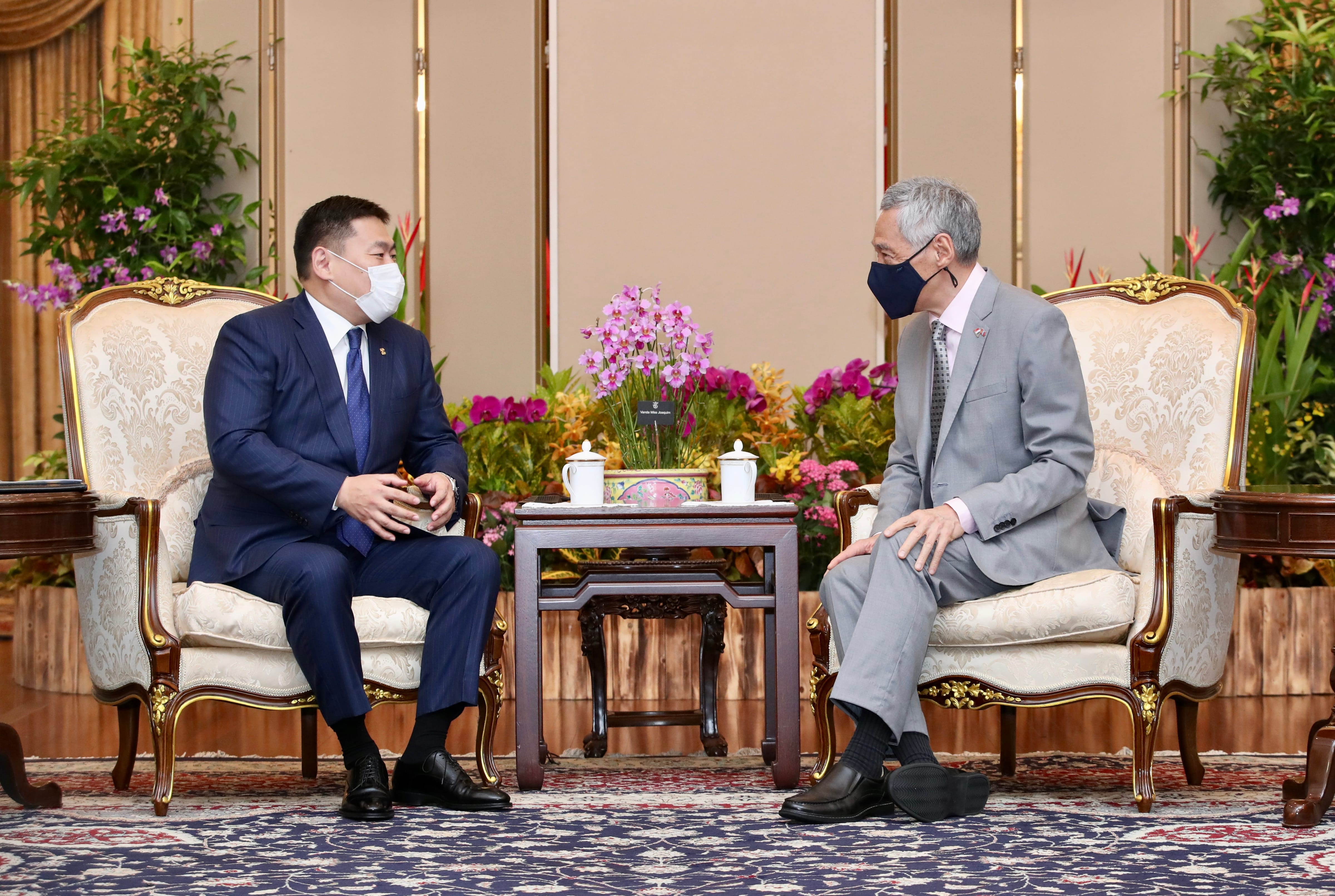 Two men wearing face masks sit in ornate chairs facing each other, a flower arrangement between them.
