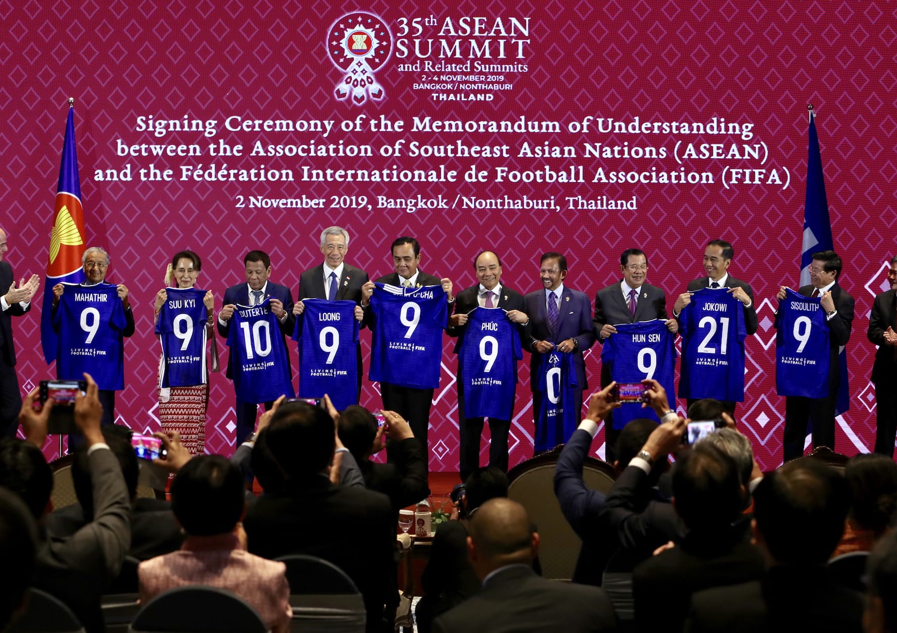 ASEAN leaders holding blue FIFA jerseys numbered 9, 10, or 21 during signing ceremony. Audience visible.