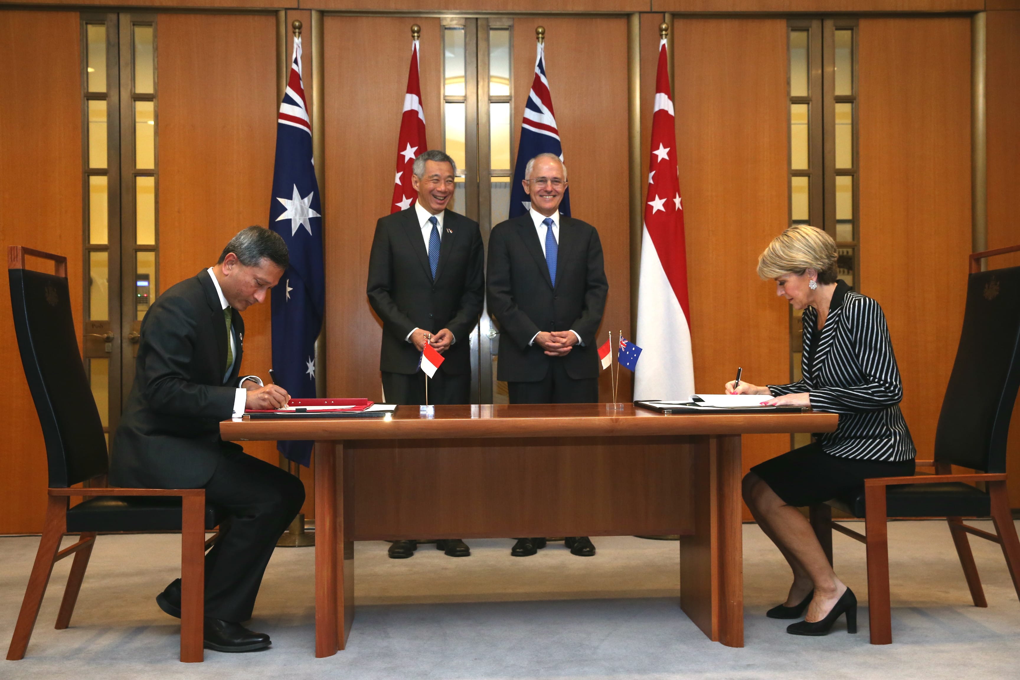 Two people sign documents at a table with flags of Australia & Singapore behind them.