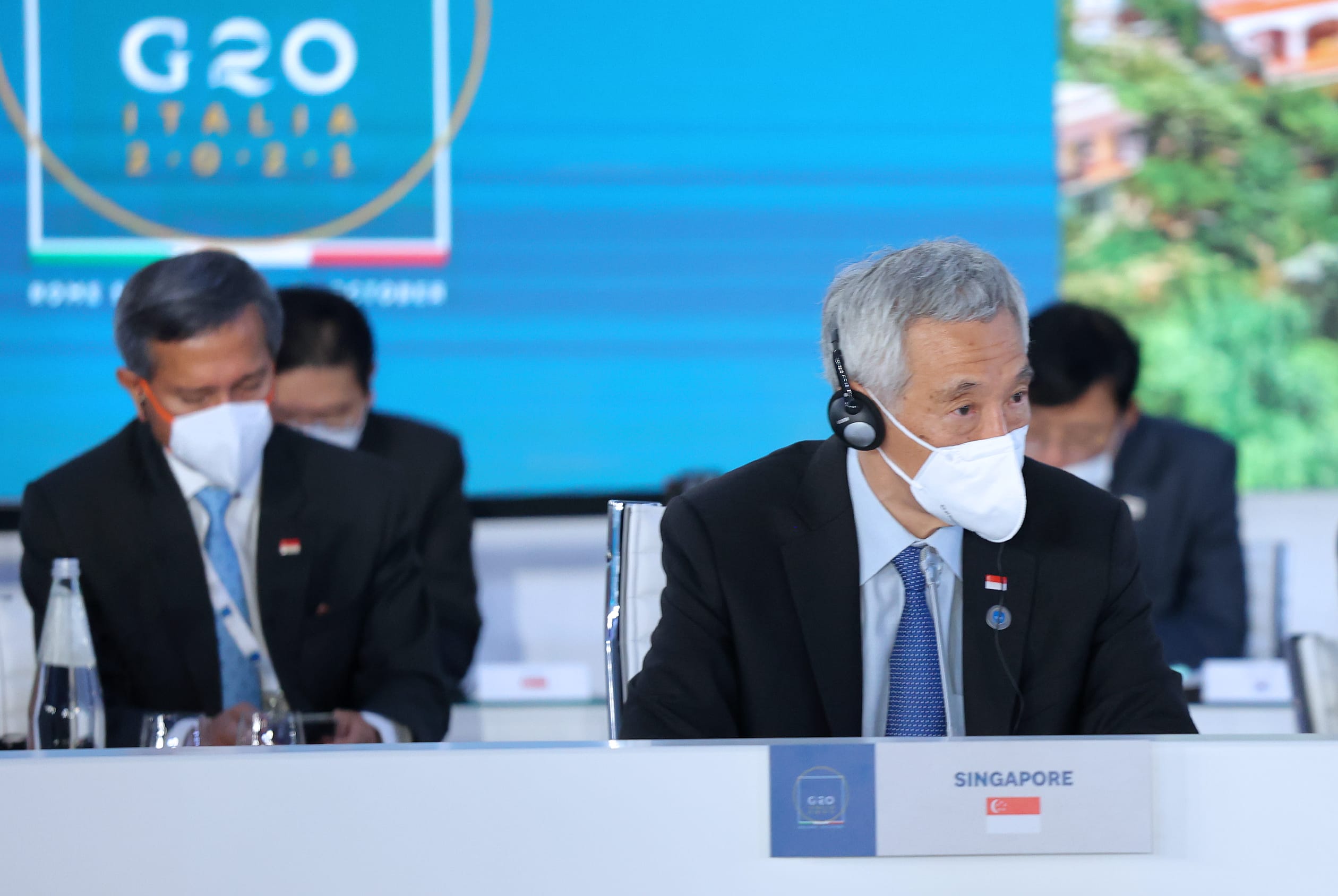 Lee Hsien Loong, with a mask and headset, sits behind a "Singapore" placard. G20 logo in background.
