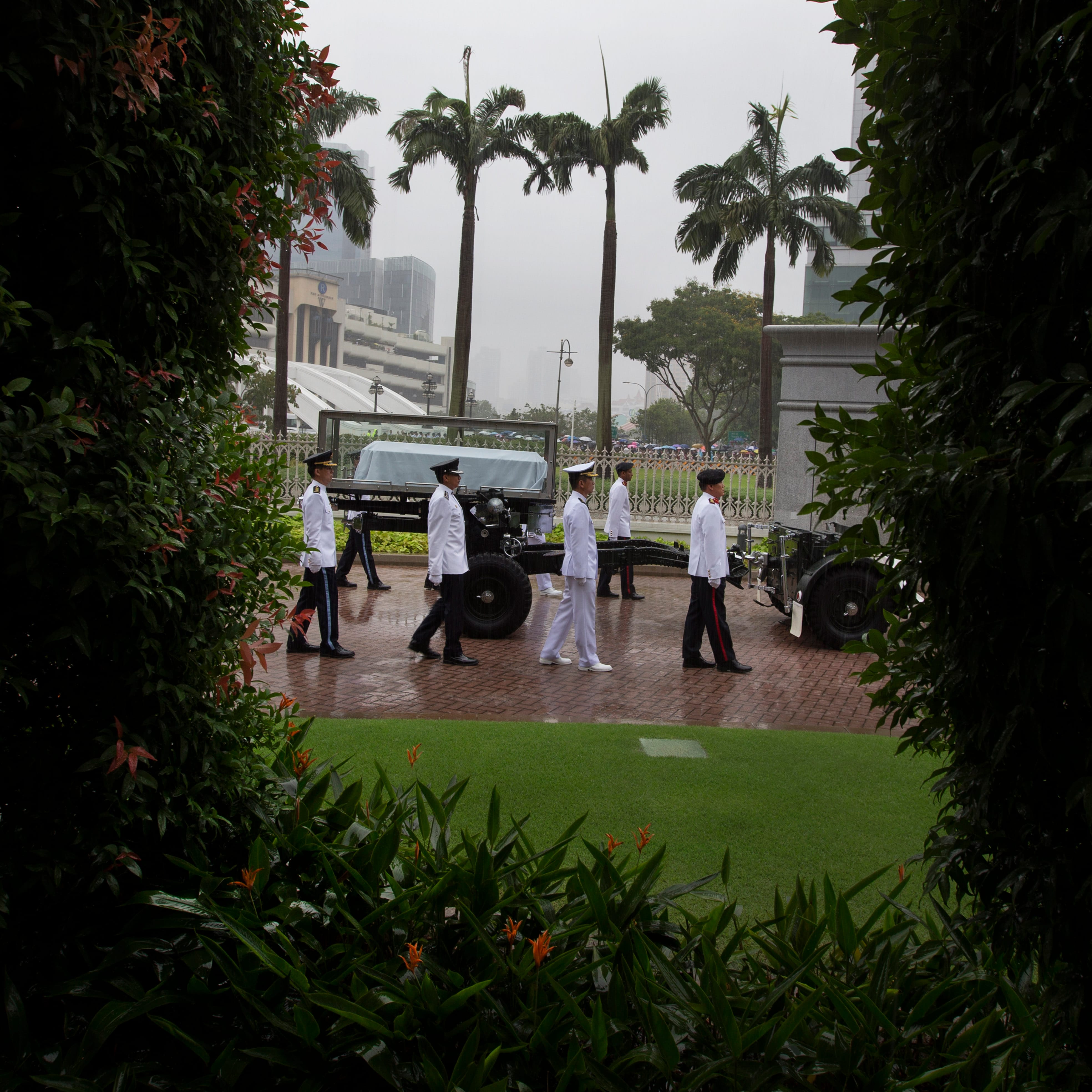 Military honor guard in white uniforms pulling a cannon with a draped coffin through rain.