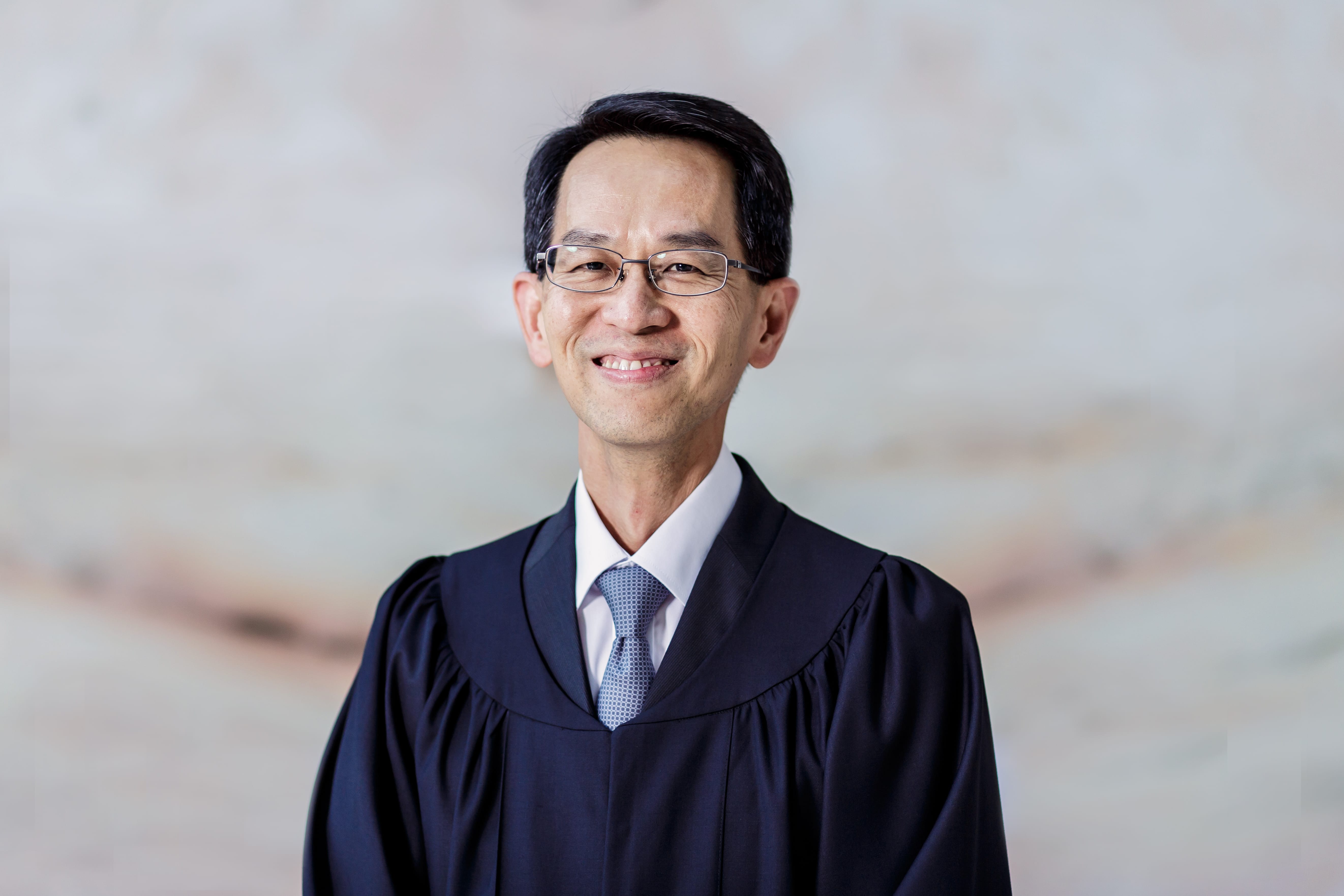 Man in black robe with glasses, white shirt, and blue tie smiles against mottled background.