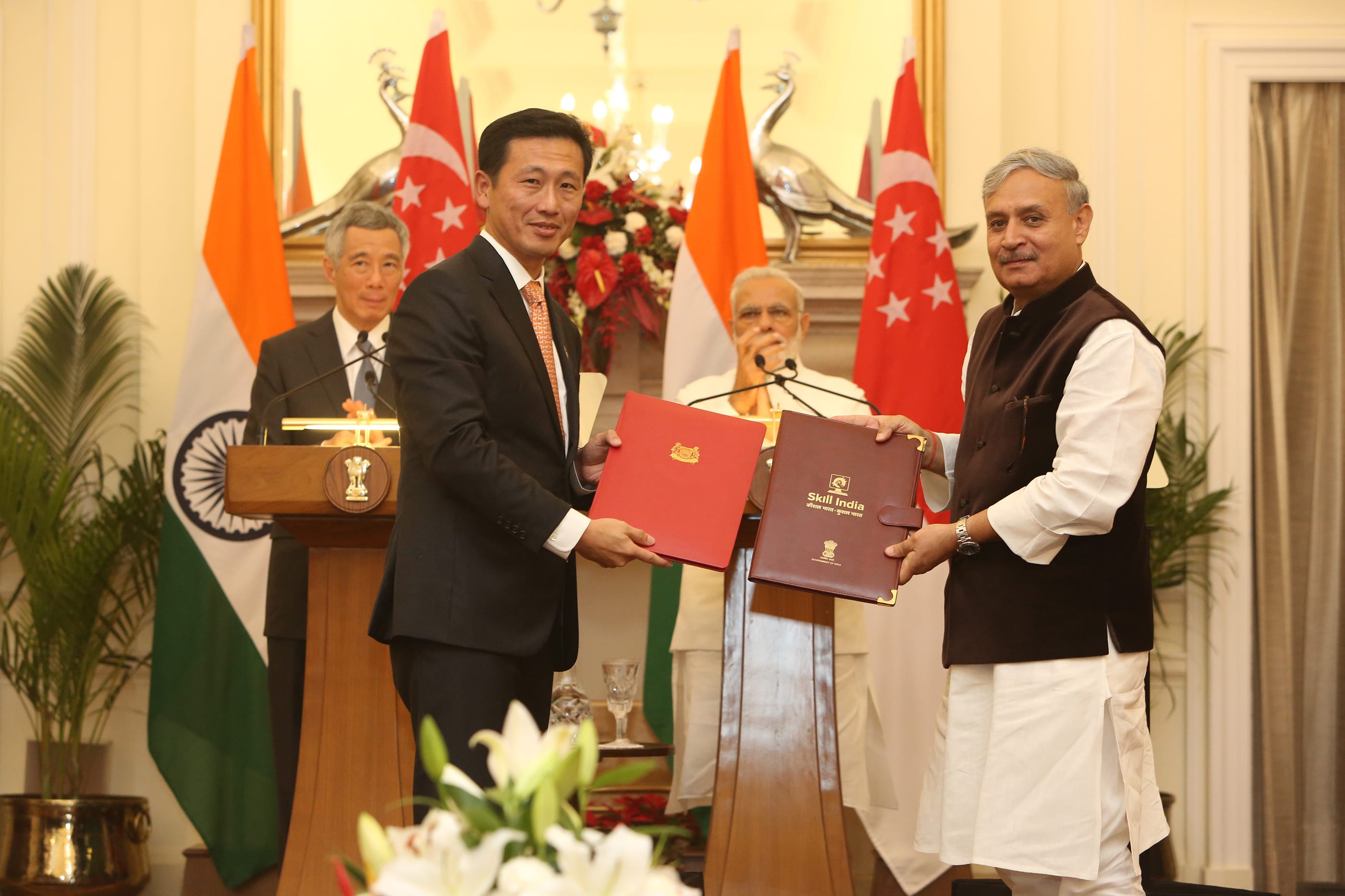 Two men exchange folders, with flags of India and Singapore behind. One folder reads "Skill India".
