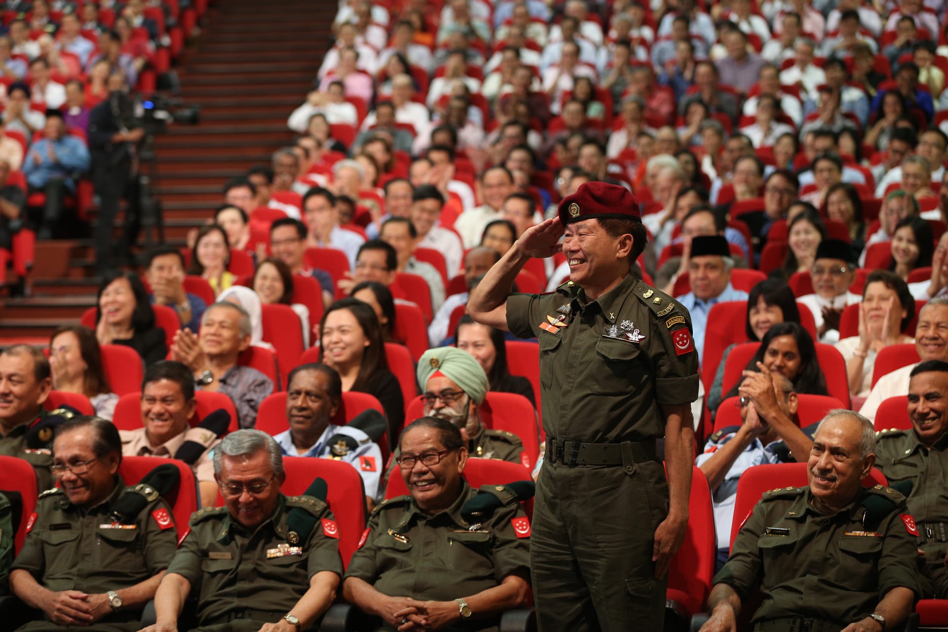 Audience in red seats; military officer in uniform saluting.