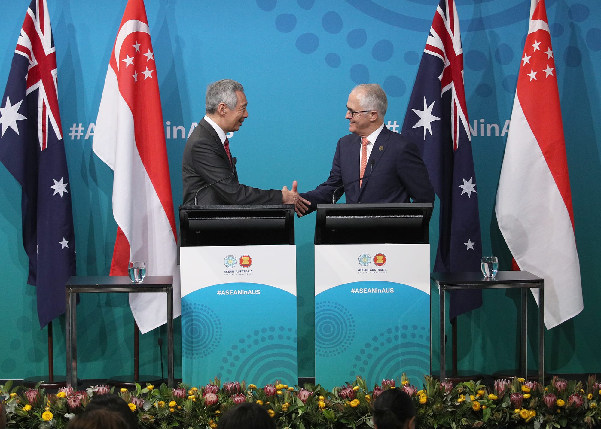 Two men in suits shake hands at podiums flanked by Australian and Singaporean flags.