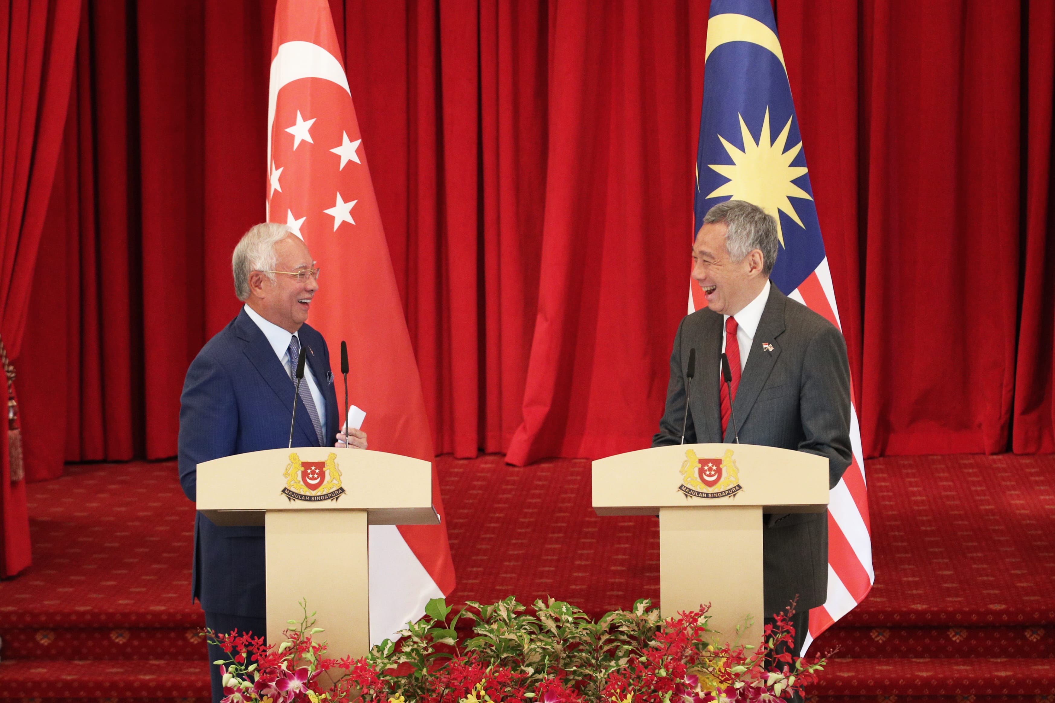 Two men at podiums with Singapore and Malaysia flags against red curtain backdrop.