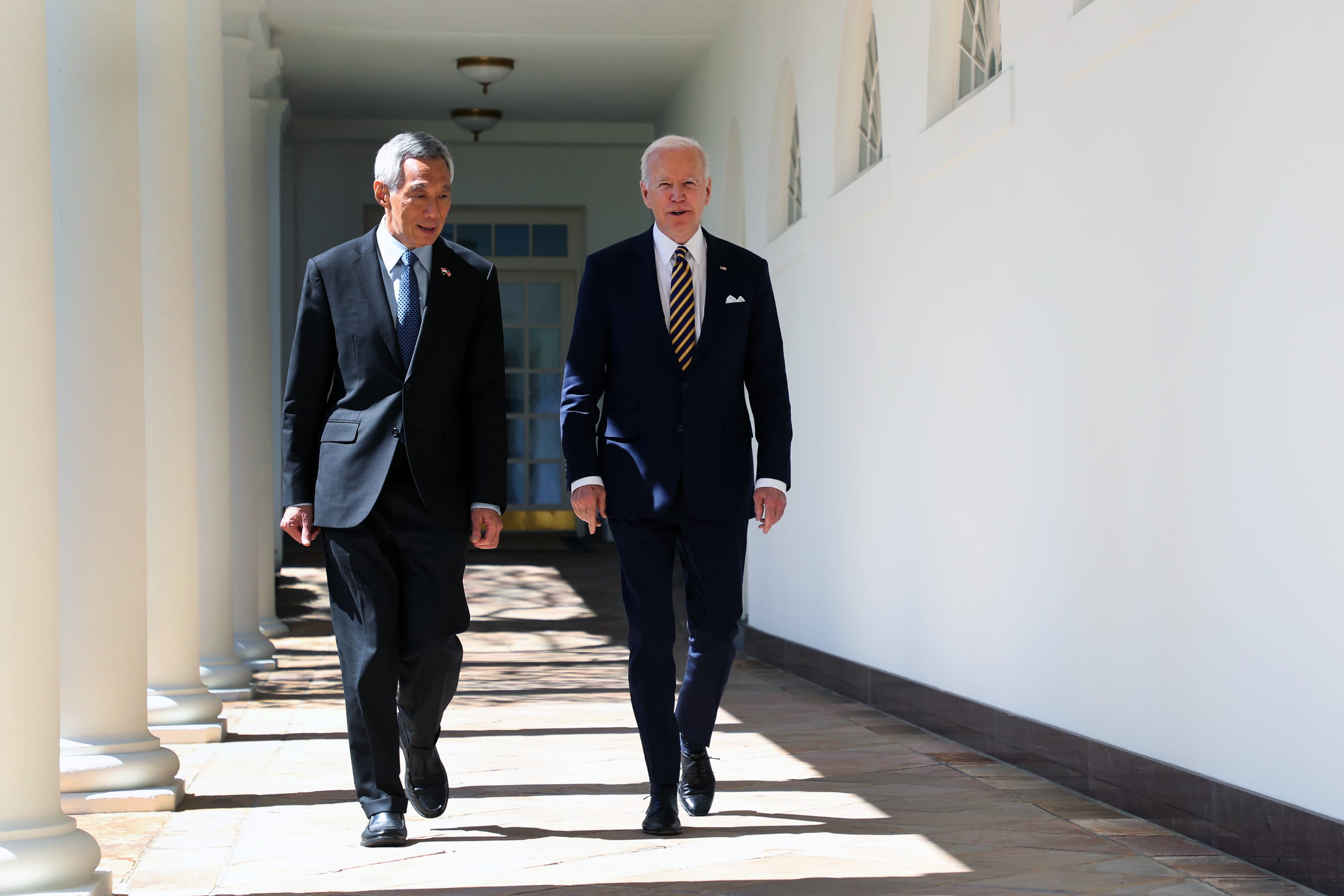 President Biden and Lee Hsien Loong walk along a colonnaded walkway. Both men are wearing dark suits.