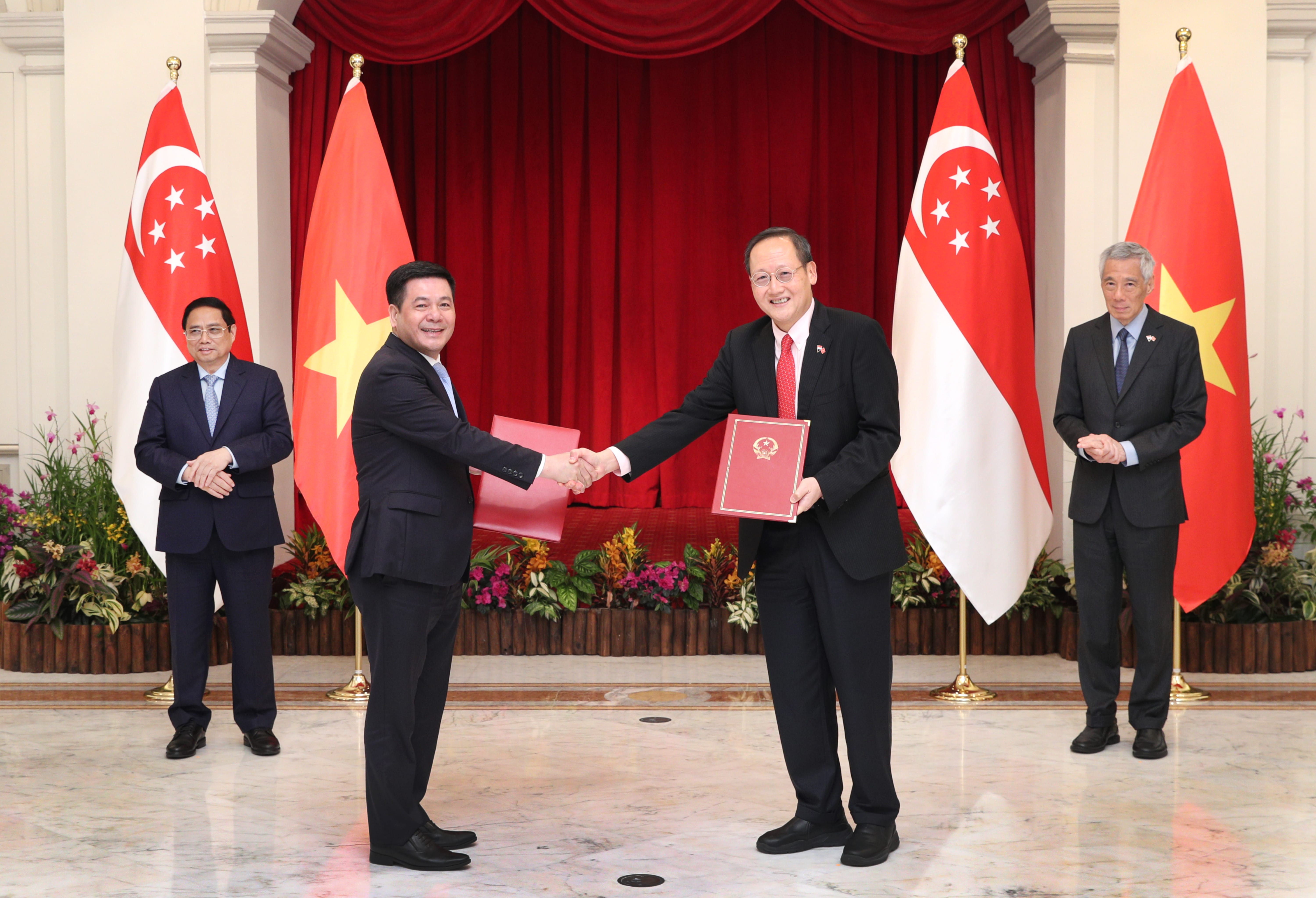 Two men in suits shake hands while holding red folders, framed by Singapore and Vietnam flags.