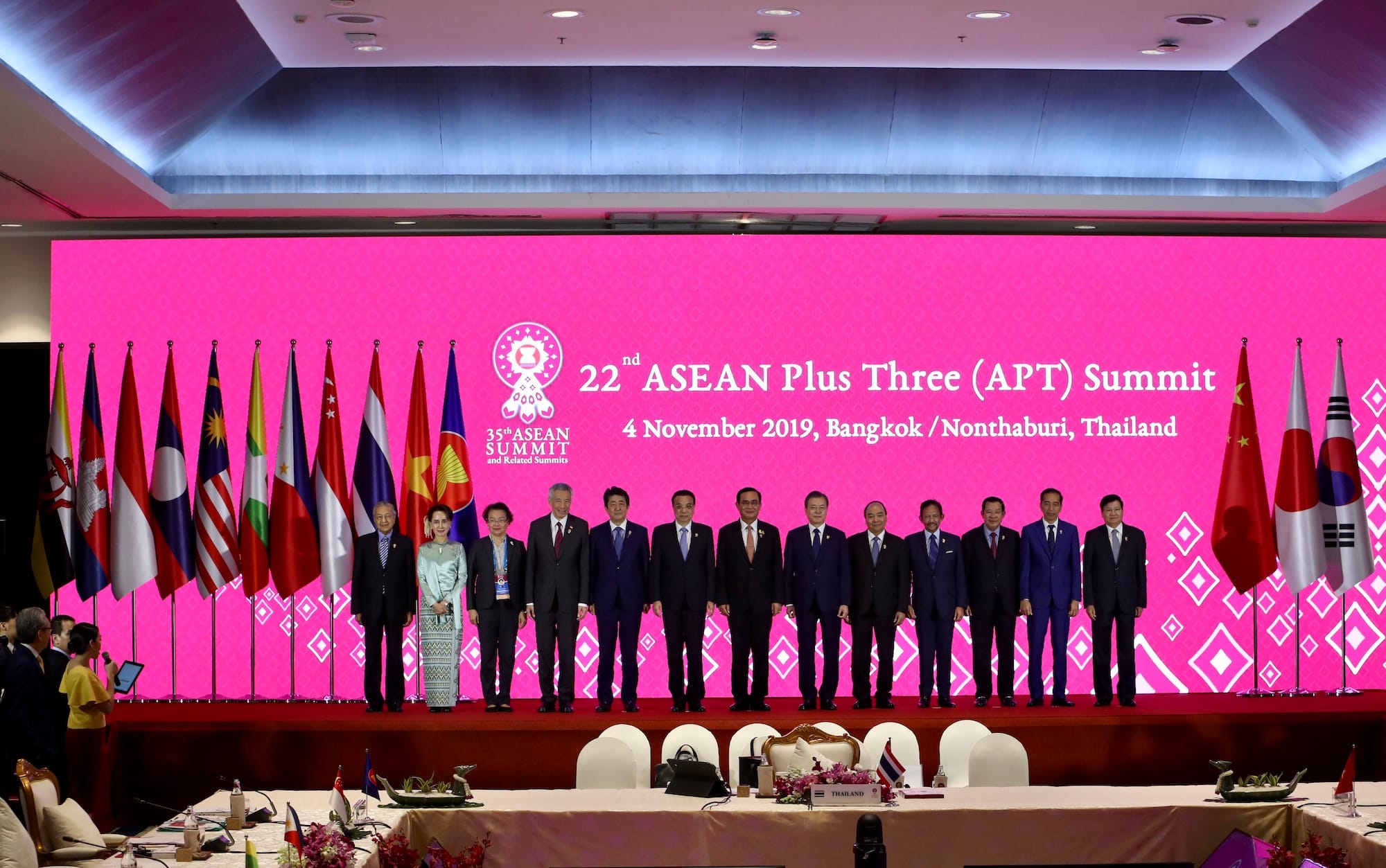 Group of leaders at 22nd ASEAN Plus Three Summit, flags behind them, pink screen backdrop.
