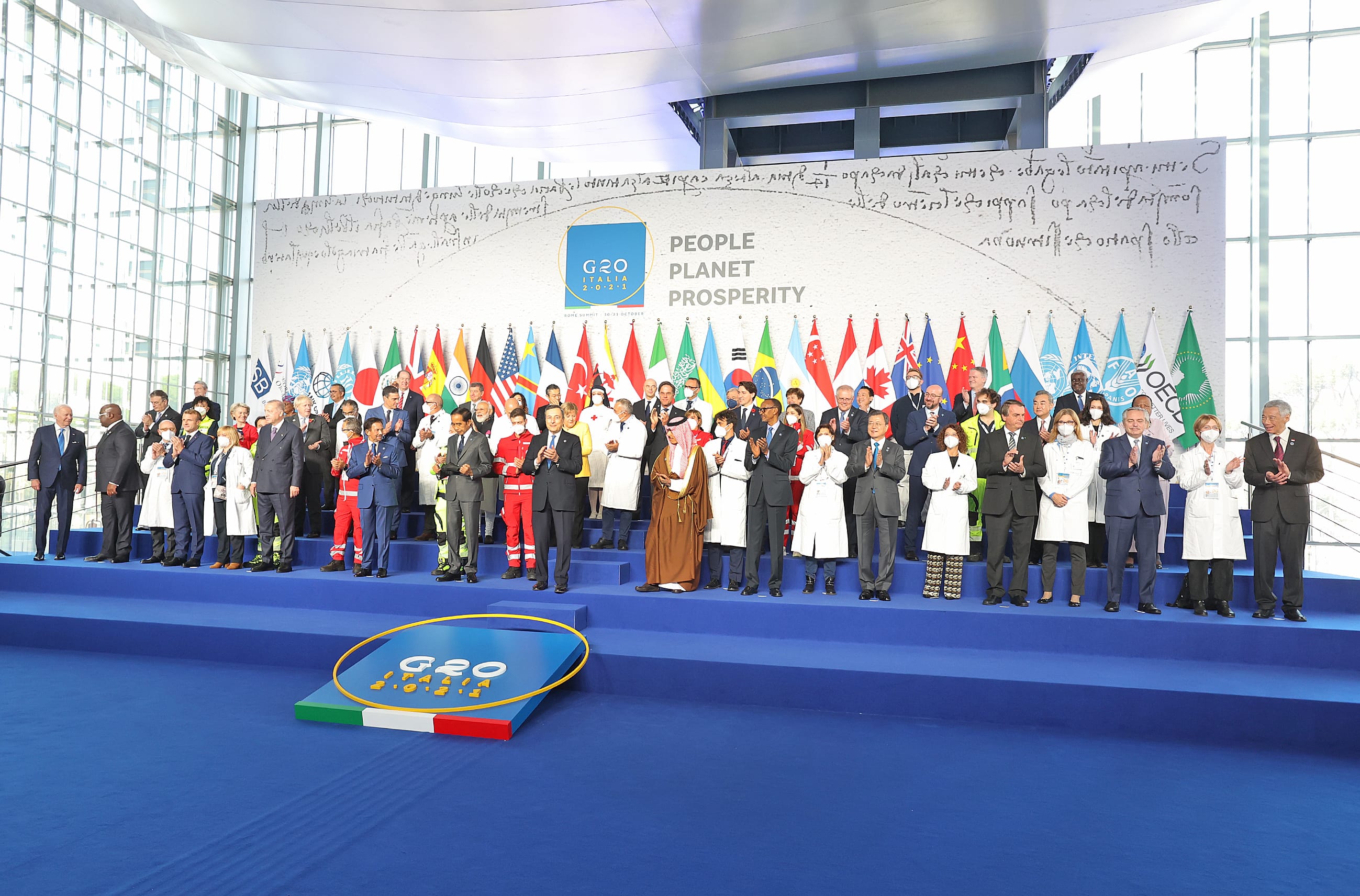 G20 Italy summit: many people stand on blue steps before flags, clapping.