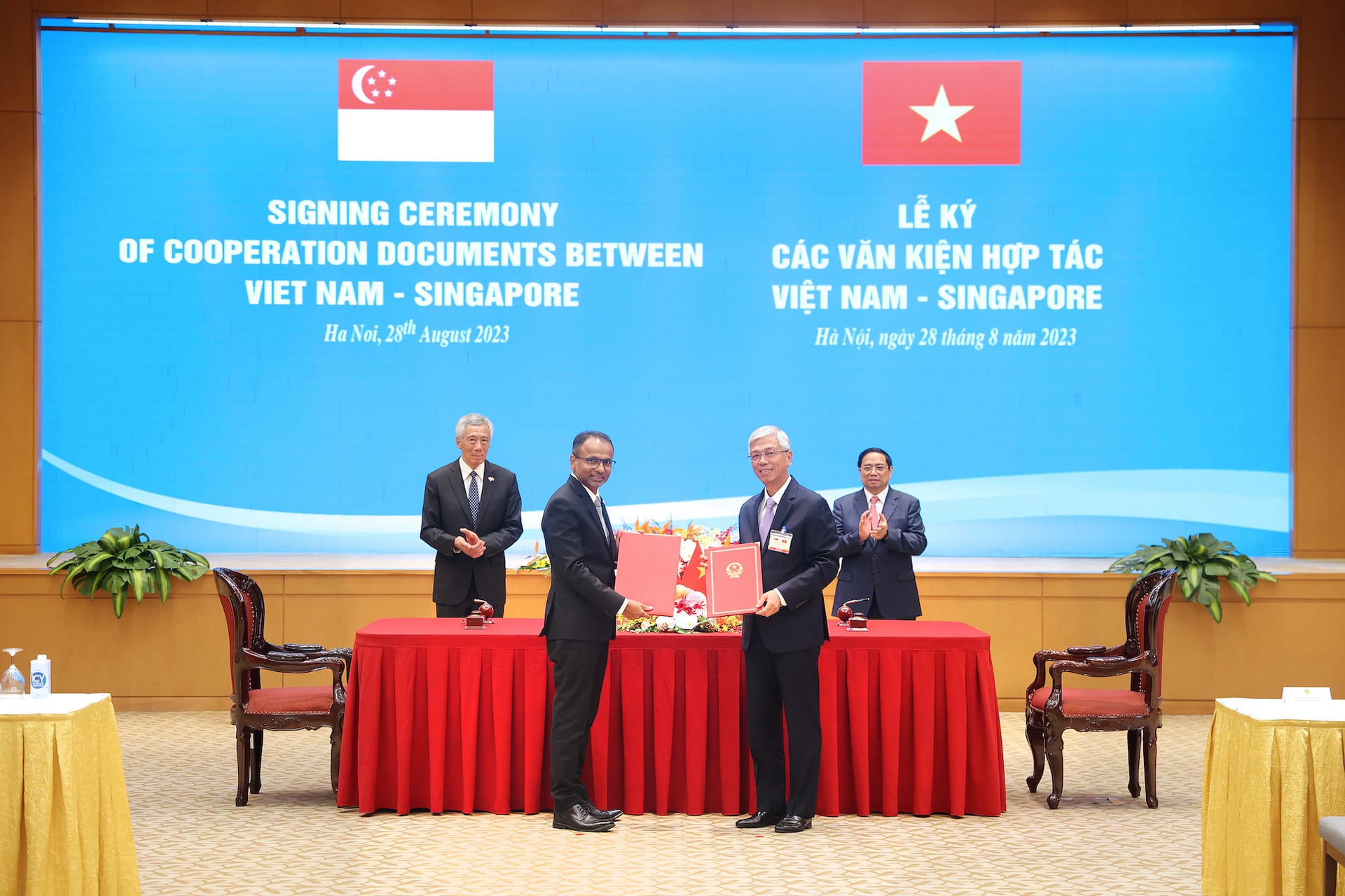 Ceremony backdrop with Singapore/Vietnam flags. Two men in suits exchange signed documents. Applause behind.