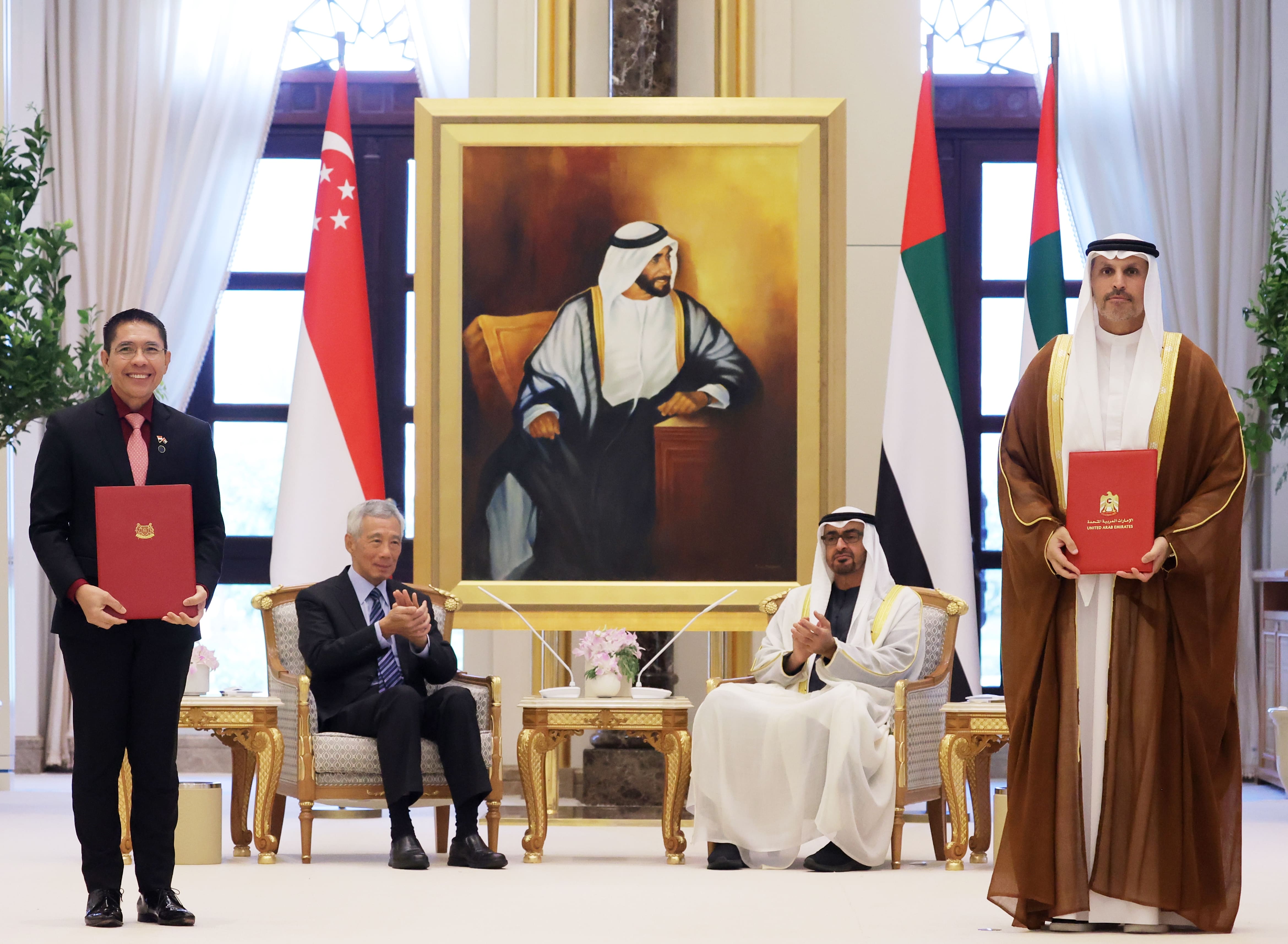 Dignitaries in formal wear with flags & a portrait. One man holds a red folder, another applauds.