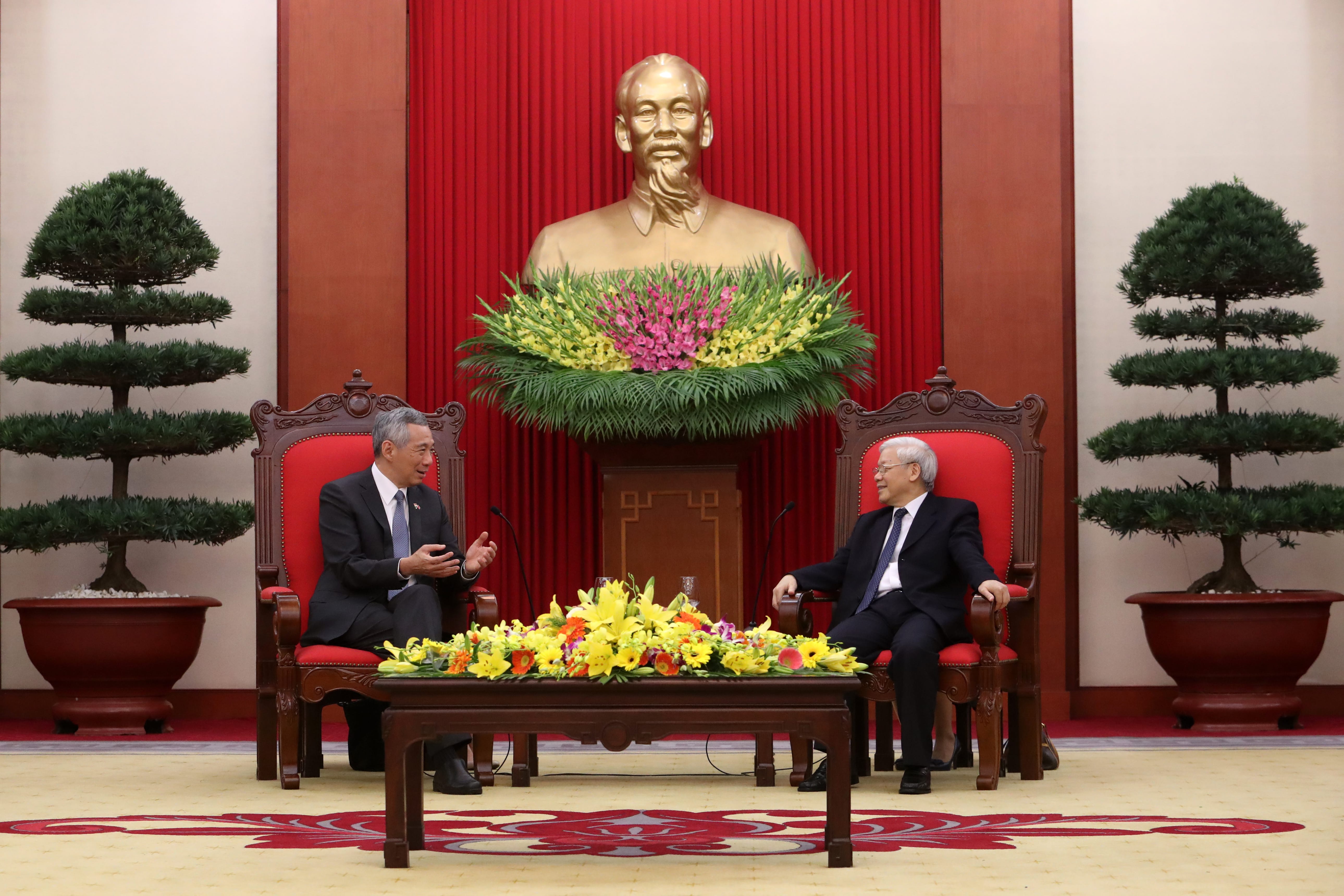 Two men in suits sit in ornate red chairs below a gold bust of Ho Chi Minh, surrounded by red drapes.
