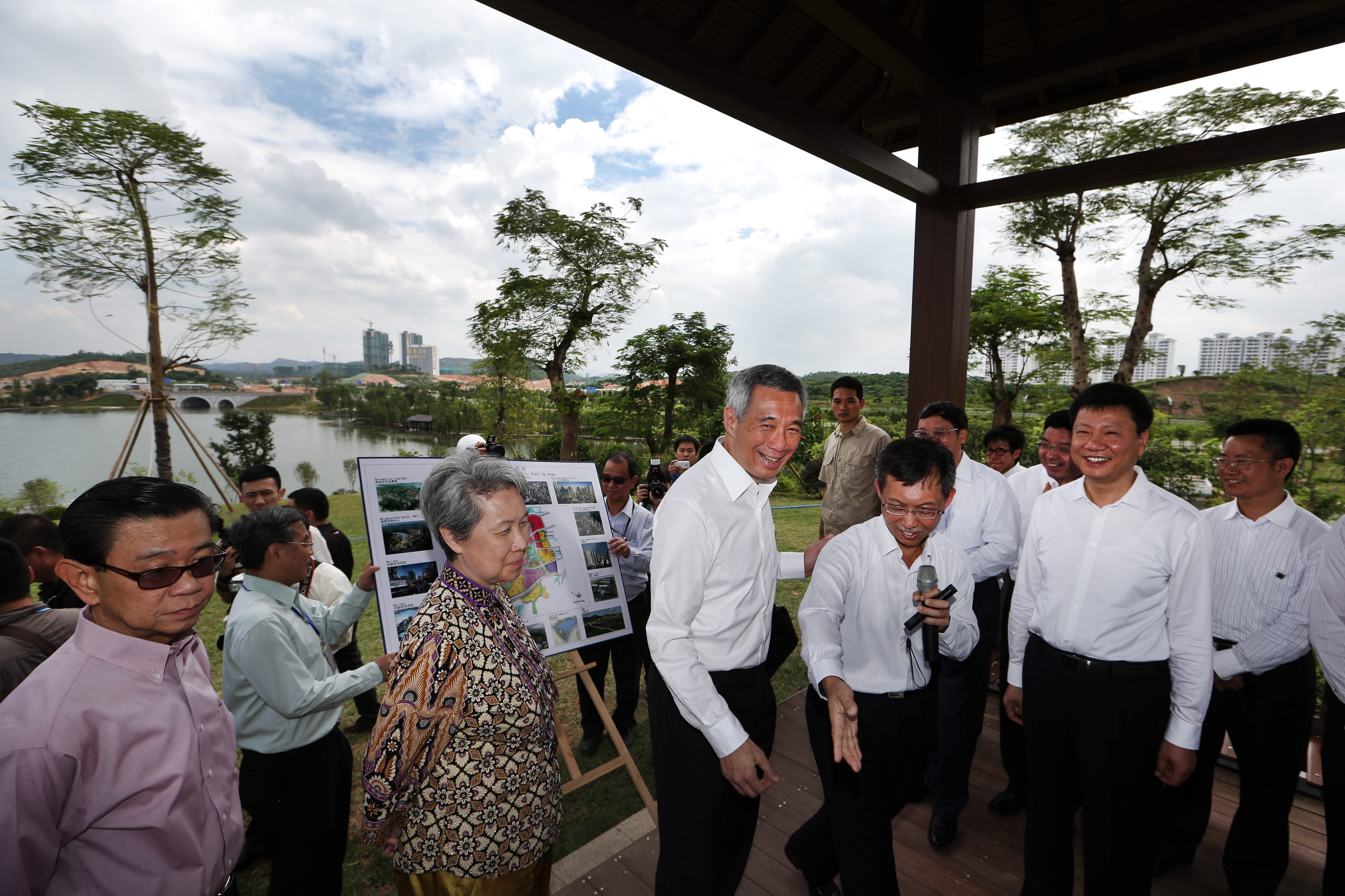 Lee Hsien Loong with group, viewing architectural plans, outdoors near a body of water.
