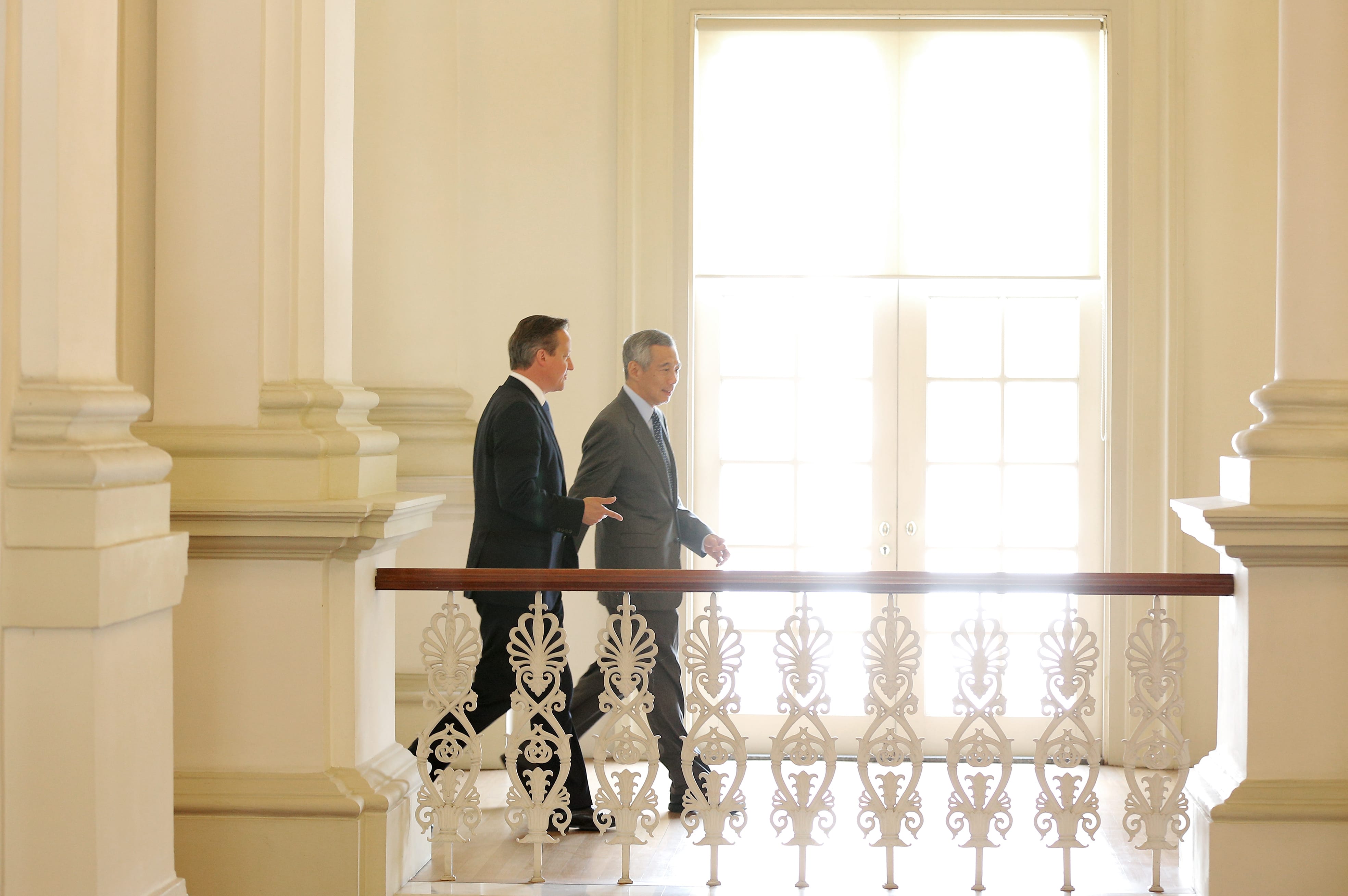 Two men in suits walk along a bright corridor behind a decorative white railing.