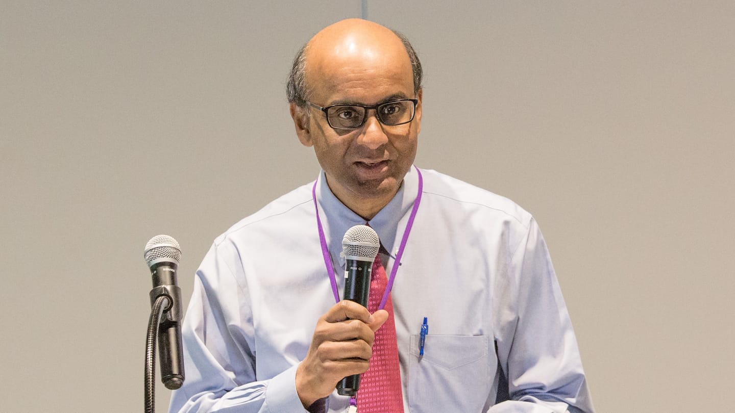 Man in glasses, light blue shirt and pink tie, holding a microphone.