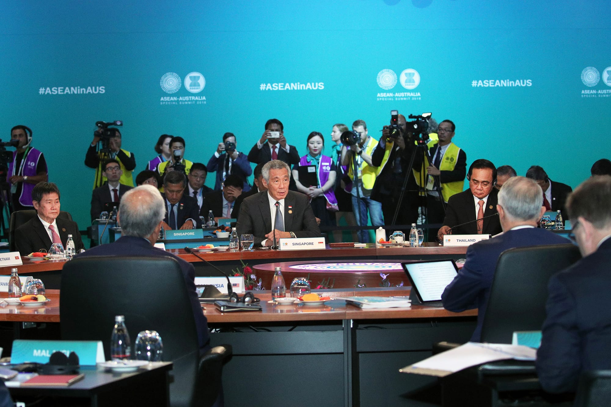 Attendees at ASEAN-Australia summit behind tables labeled "Singapore," "Thailand" and "Malaysia". Media in the background.
