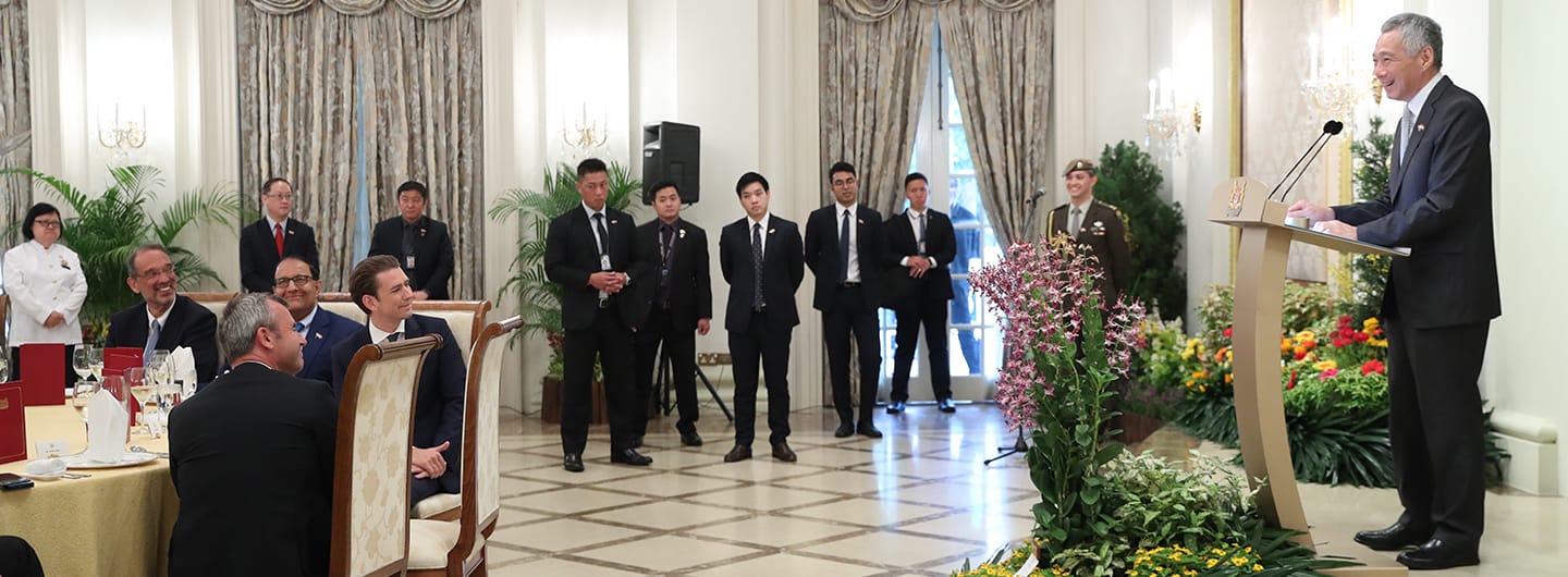 Lee Hsien Loong at podium with guards, attendees, and table setting in an ornate hall.