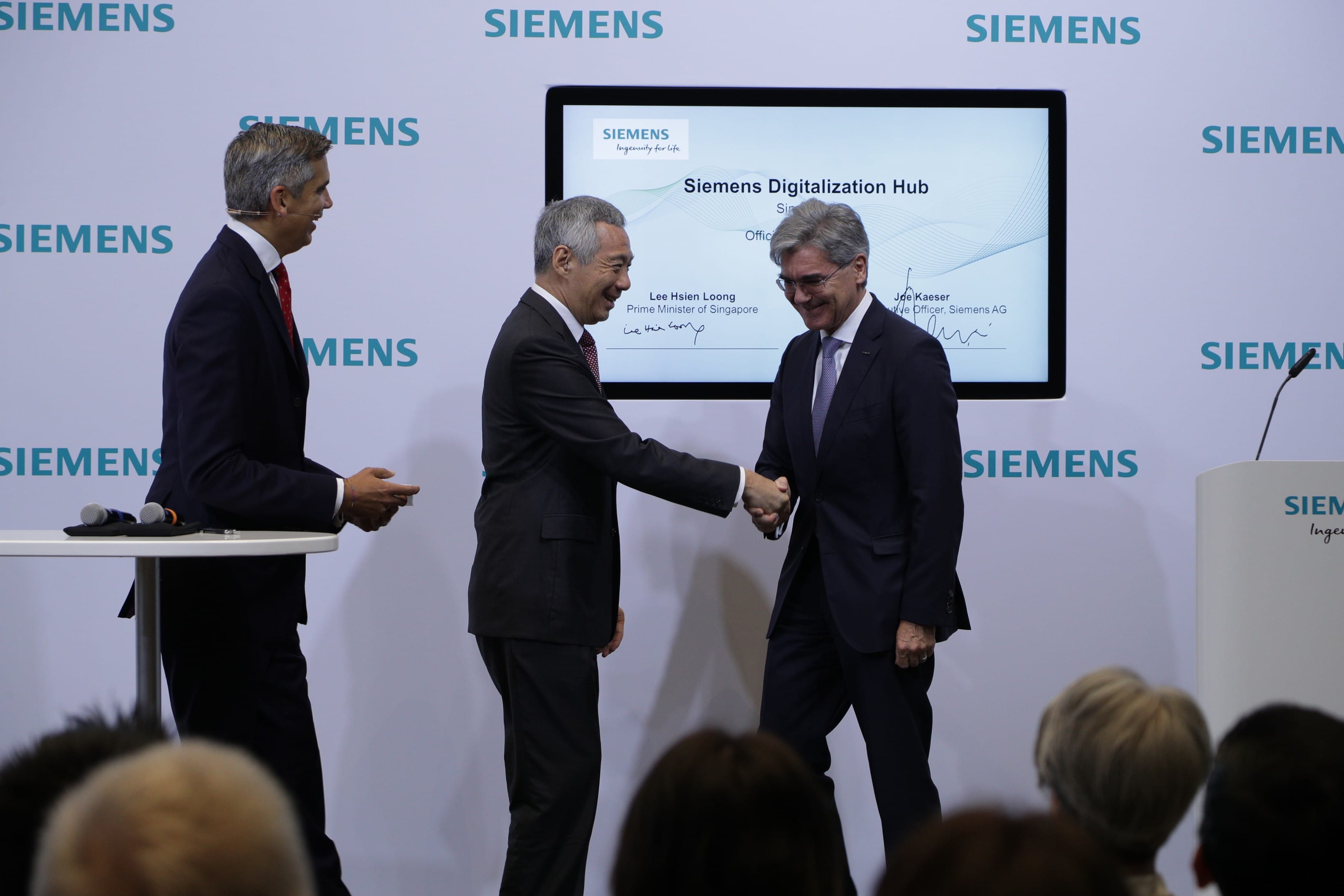 Two men in suits shake hands in front of a "Siemens Digitalization Hub" display. A third man is beside them.