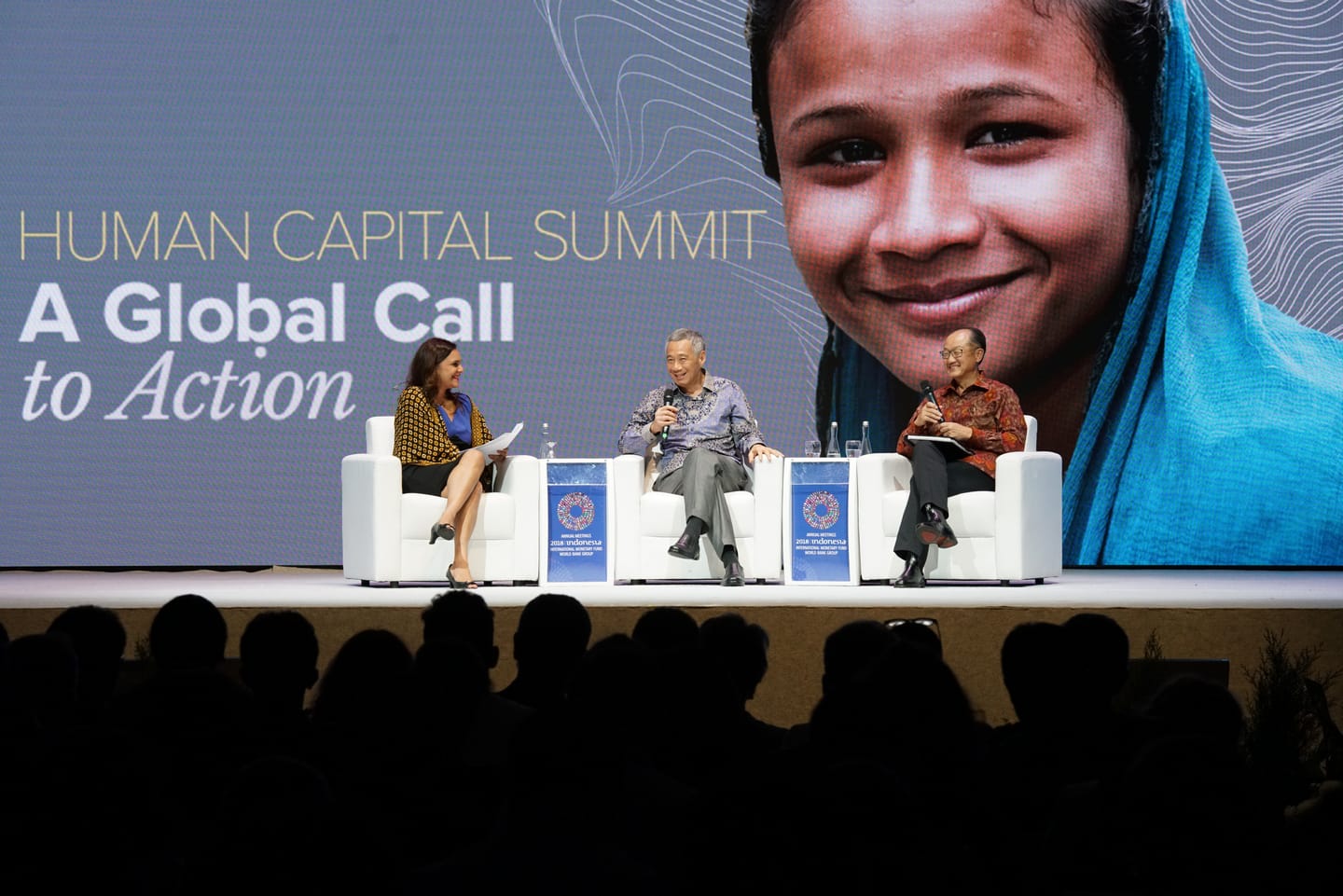 Three people on stage at "Human Capital Summit" with a girl's image as a backdrop. Crowd visible.
