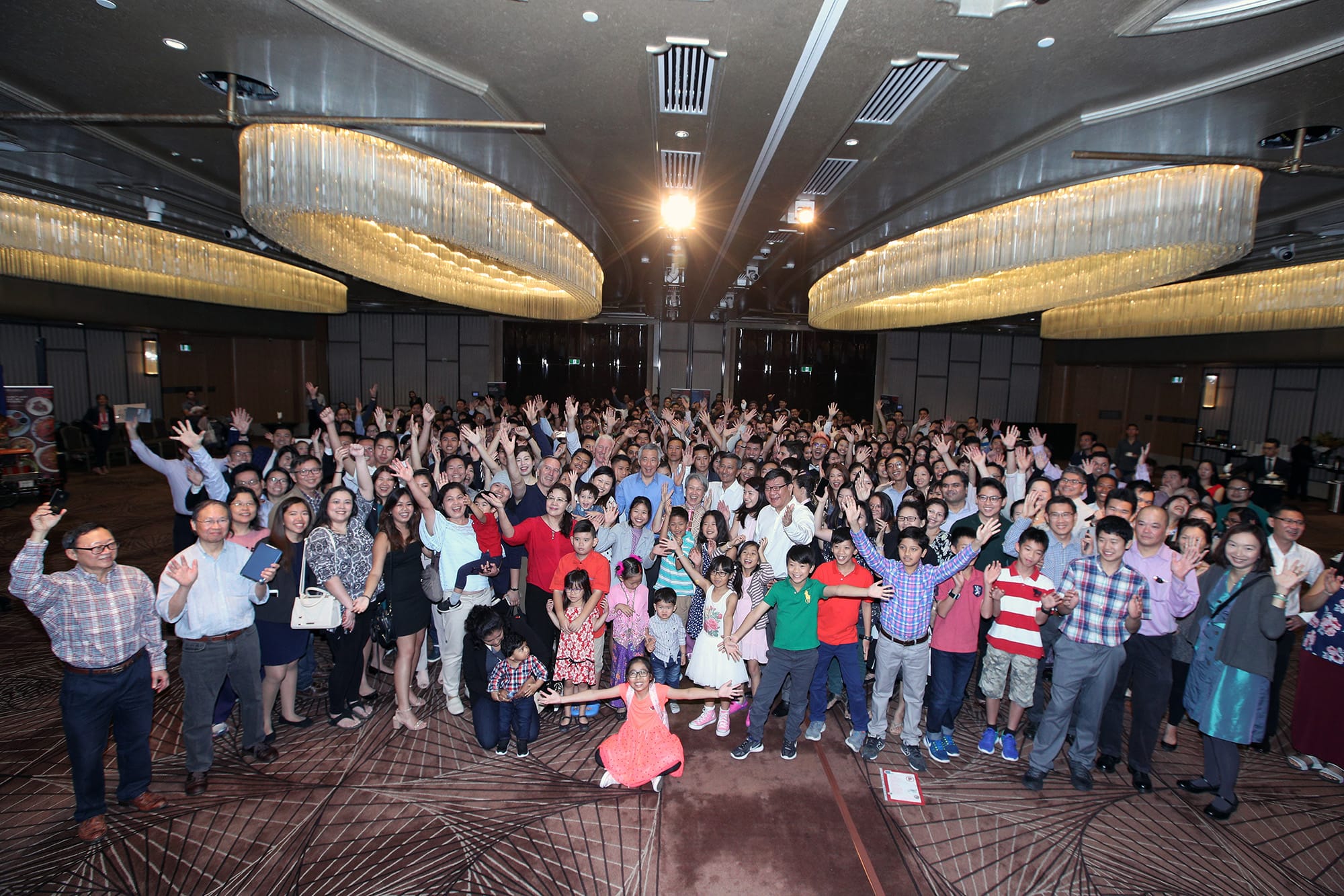 Large group of people with hands raised indoors under large chandeliers.