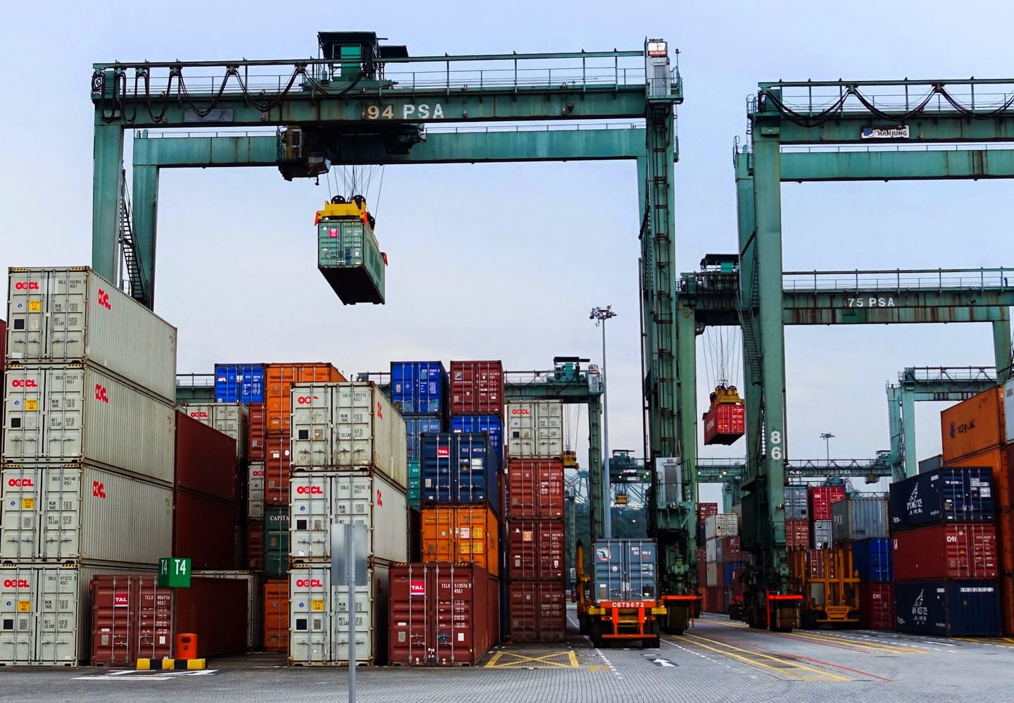 Shipping yard with cranes lifting OOCL and TAL containers stacked on the ground.
