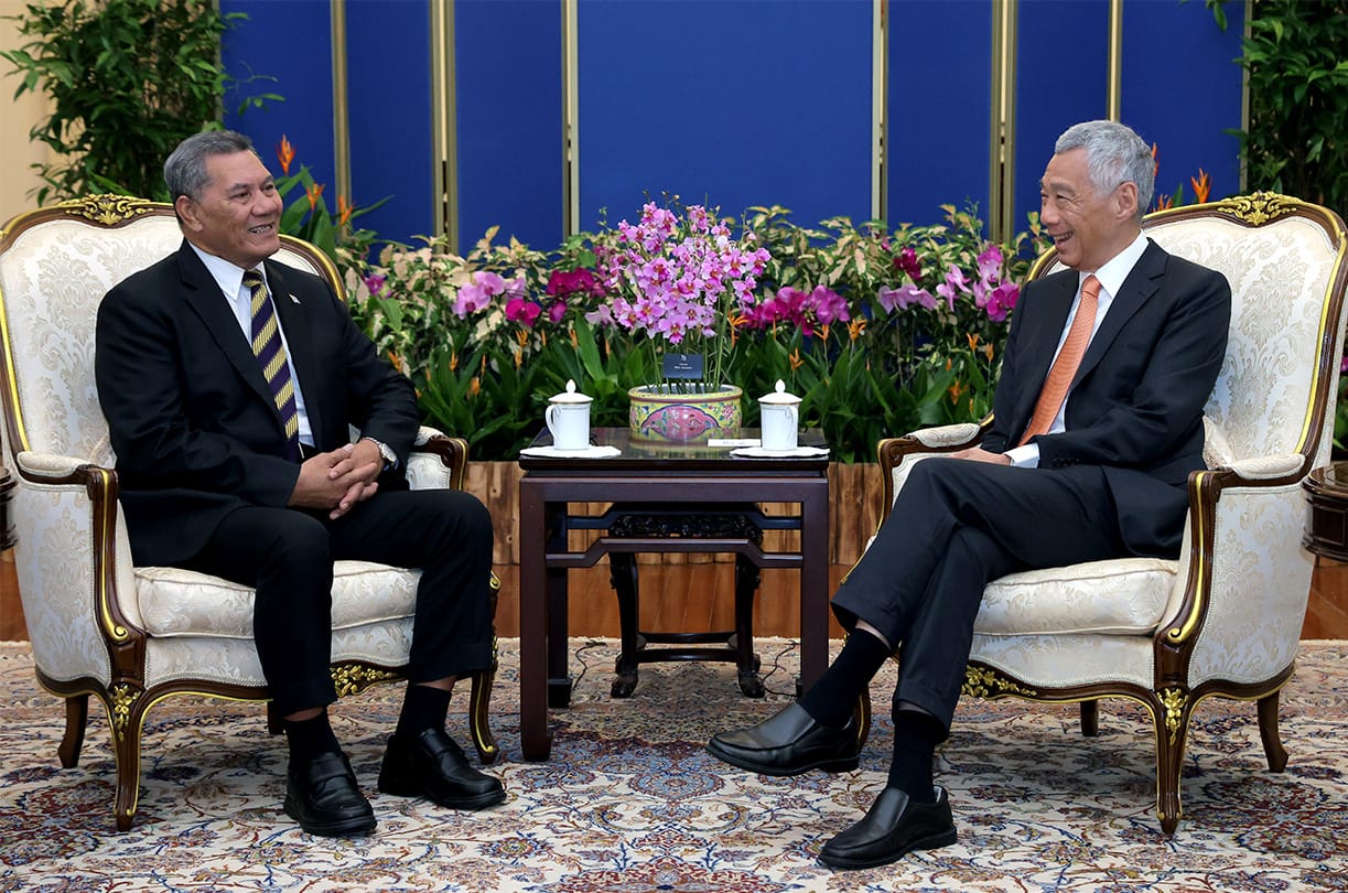 Two men in suits sit in ornate chairs, facing each other with a small table between them.
