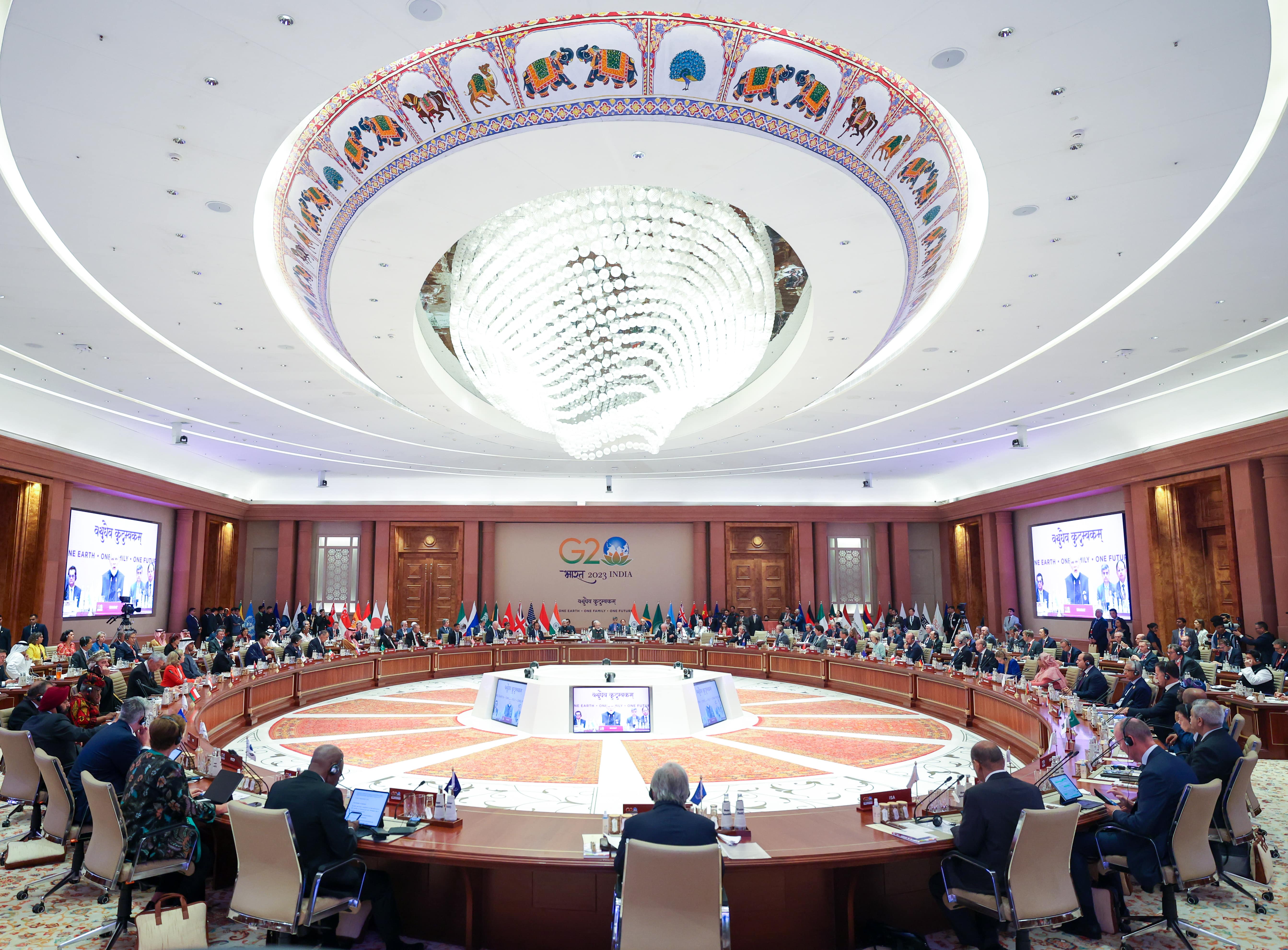 G20 summit: delegates seated around a large circular table with country flags. Overhead is a chandelier.