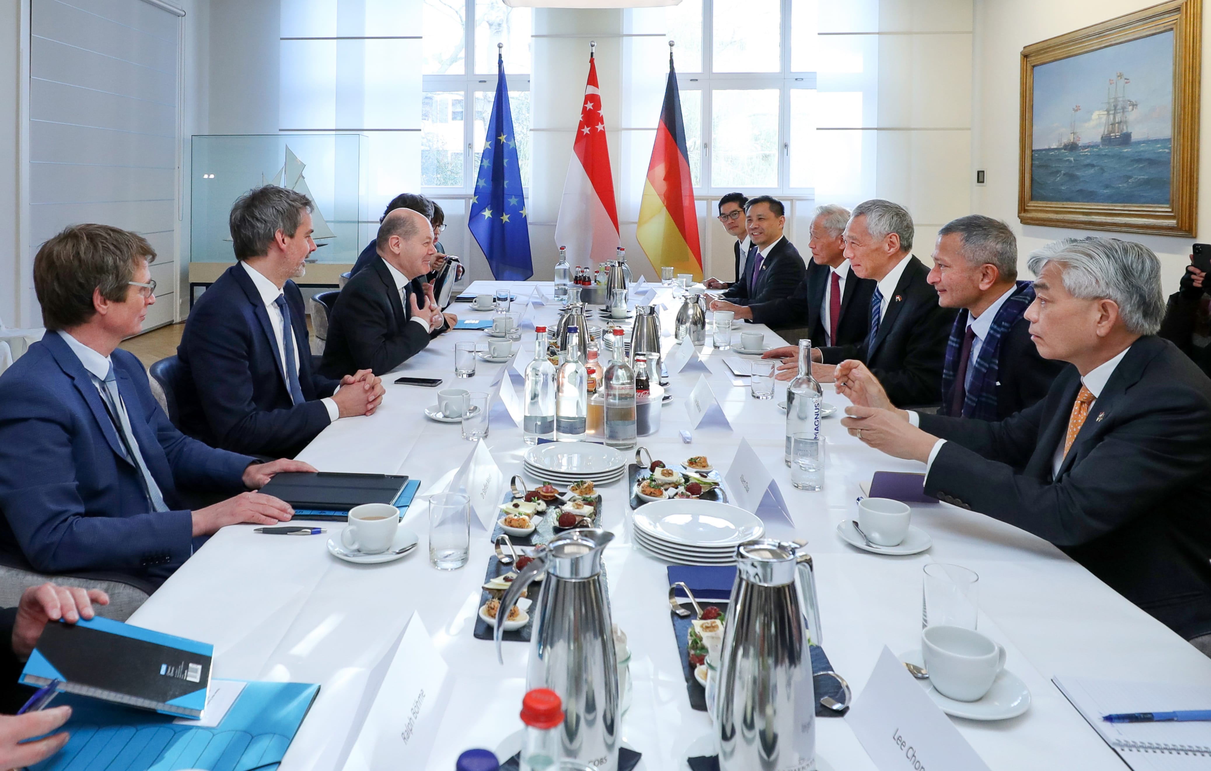 People at a table with EU, Singapore, and German flags. Table has food, drinks, and place cards.