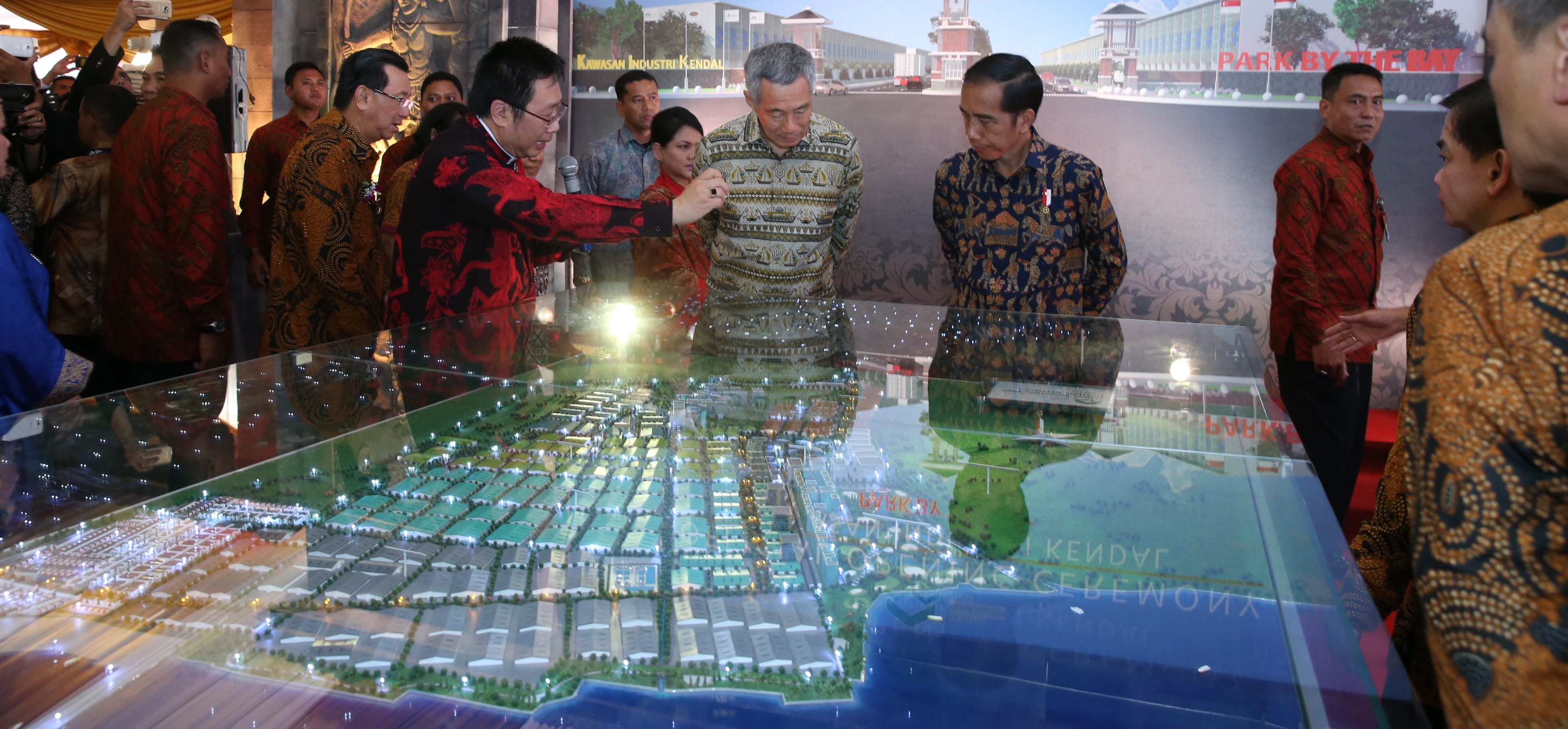 People examine a scale model city layout under glass, with an industrial park backdrop.