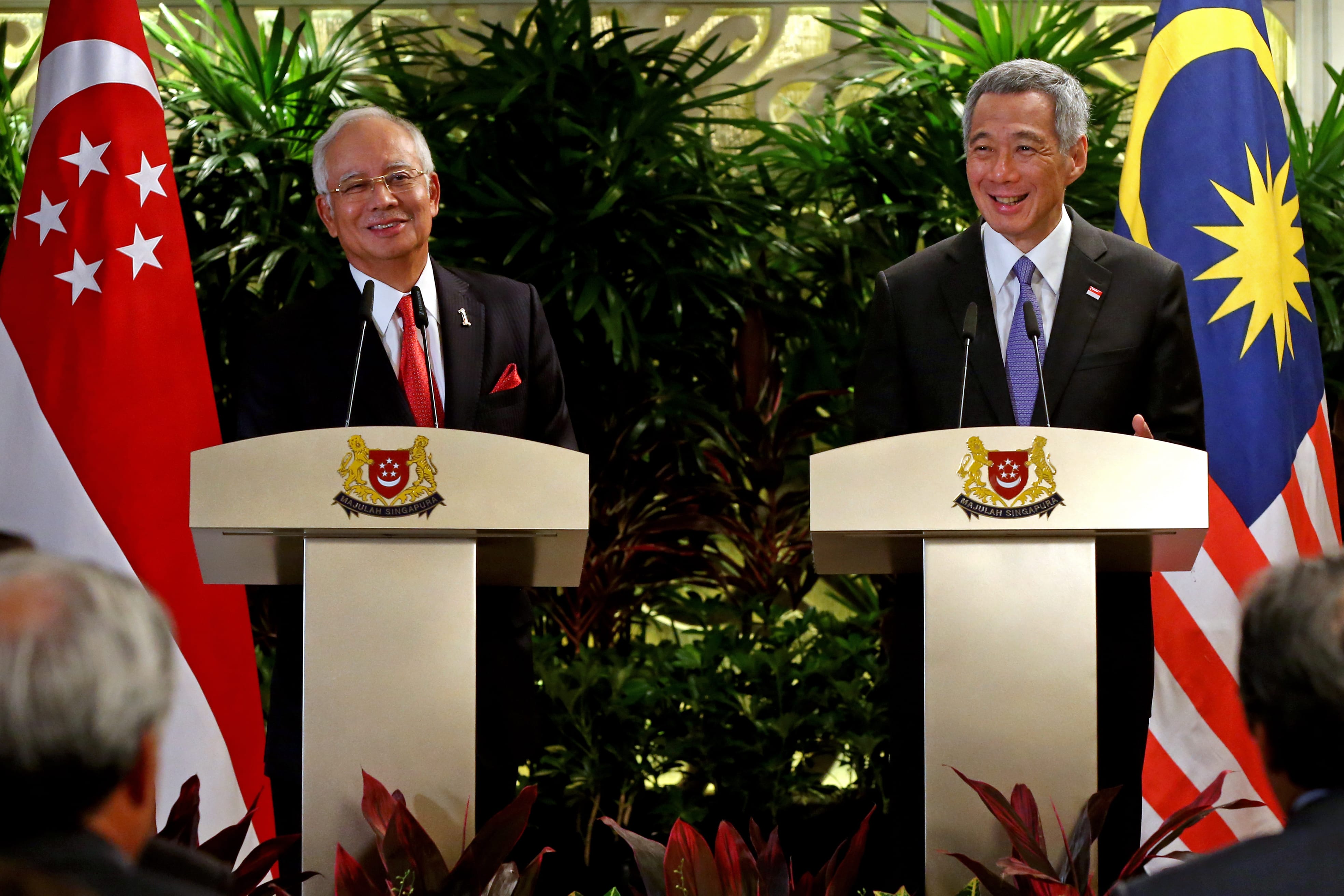 Two men at podiums with Singapore and Malaysia flags behind them.
