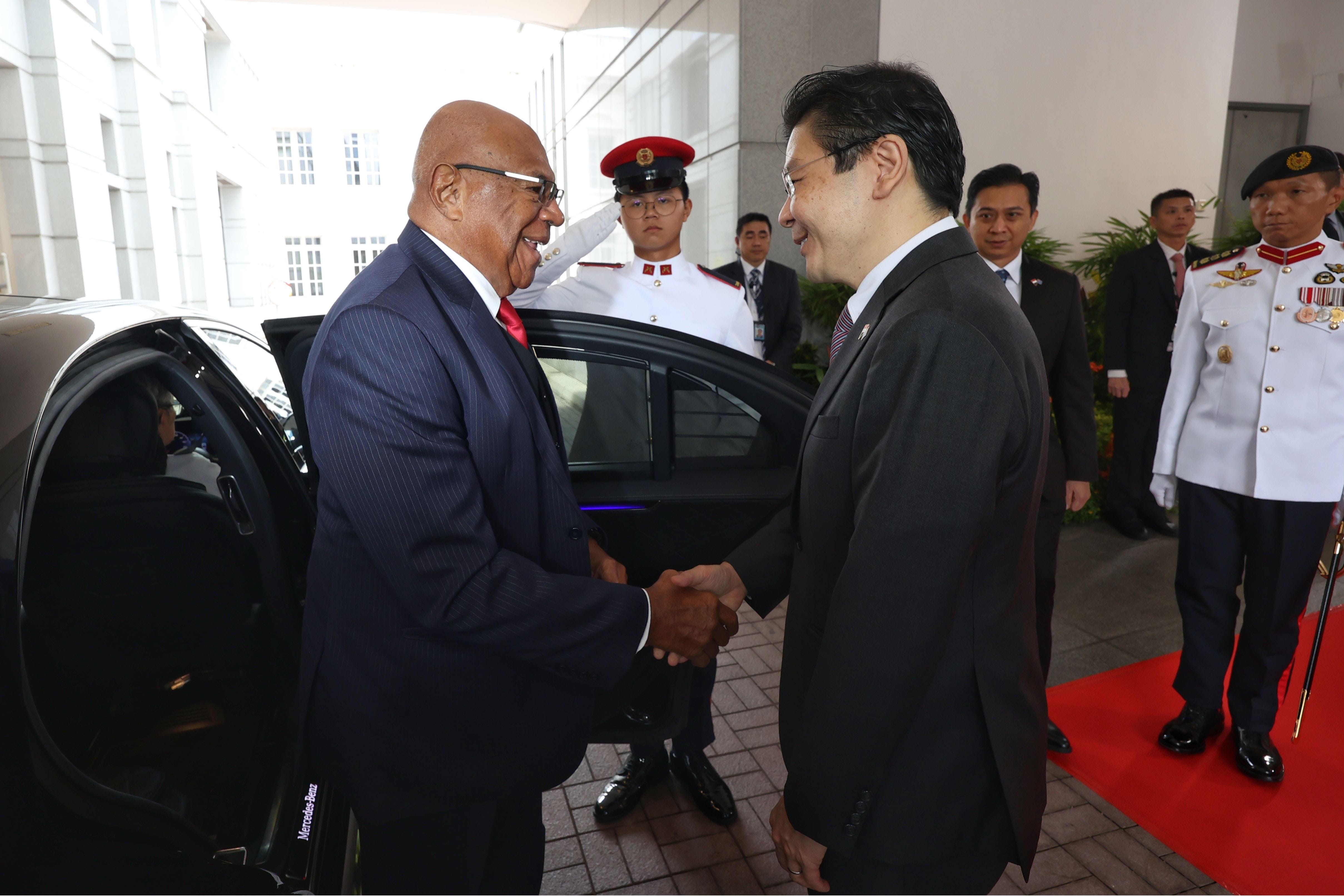 Two men in suits shaking hands by a Mercedes-Benz car, guards in dress uniforms standing by.