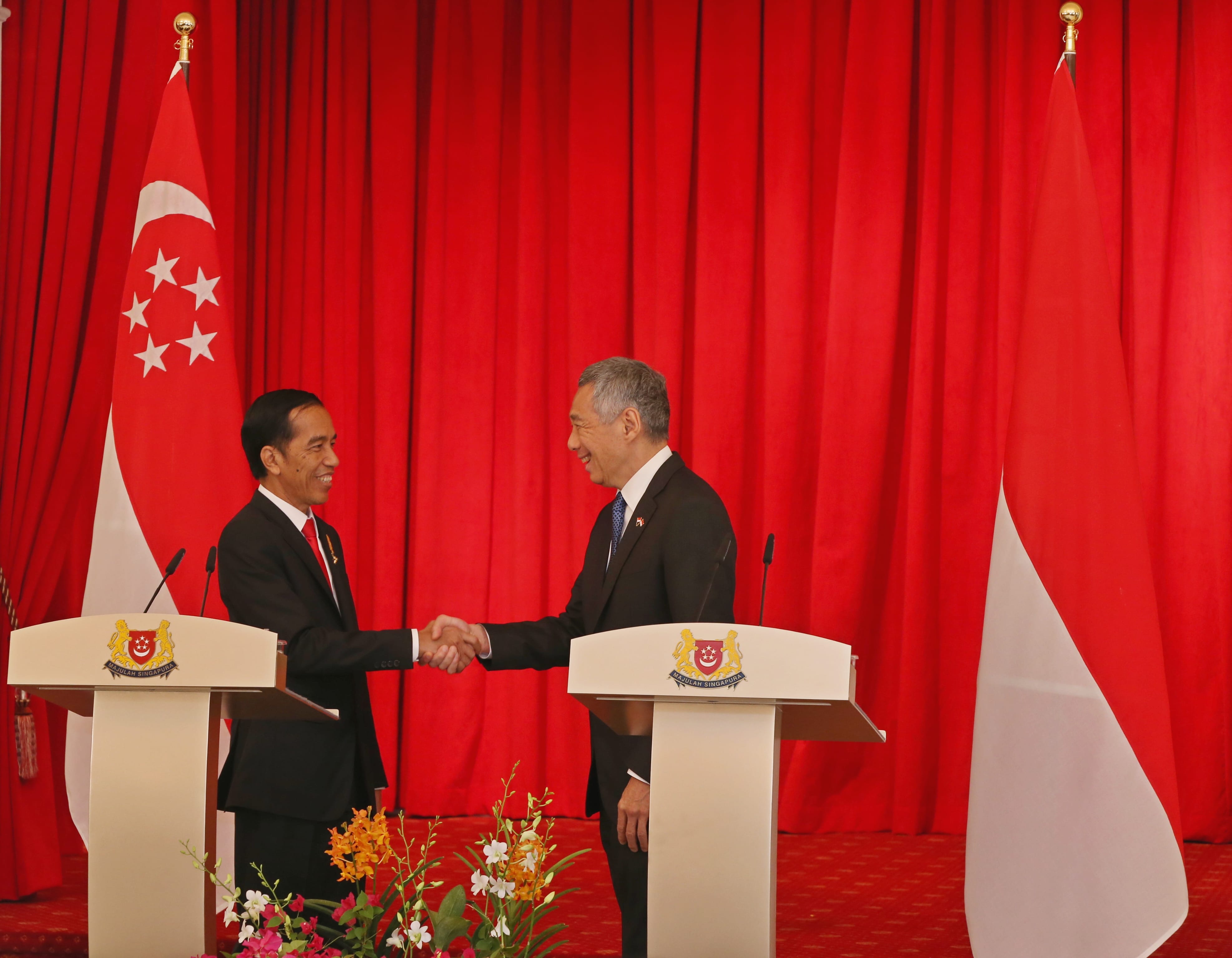 Two men in suits shake hands before Singapore and Indonesia flags on a red curtain backdrop.