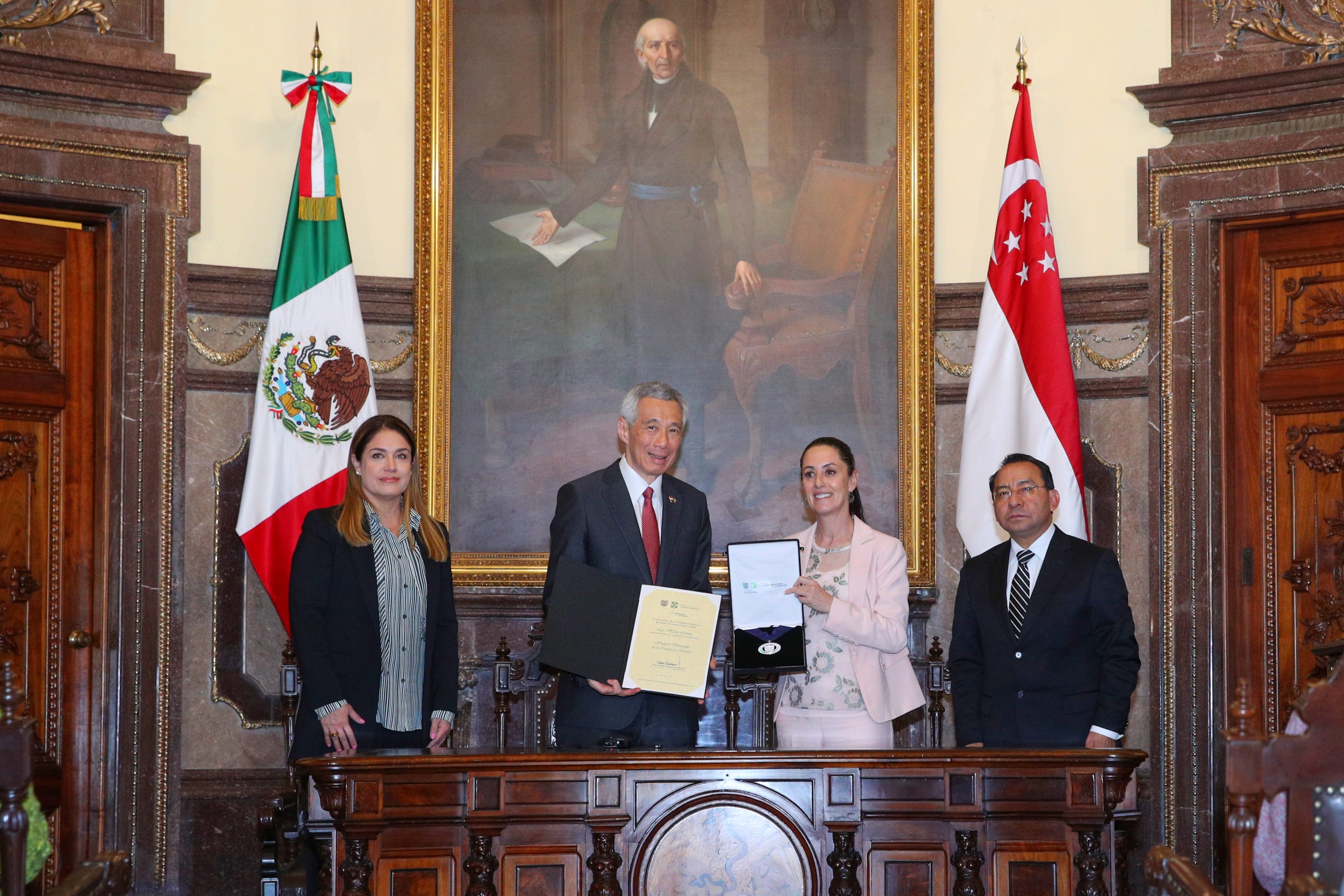 Four people behind a wooden table with Mexican and Singapore flags displayed.