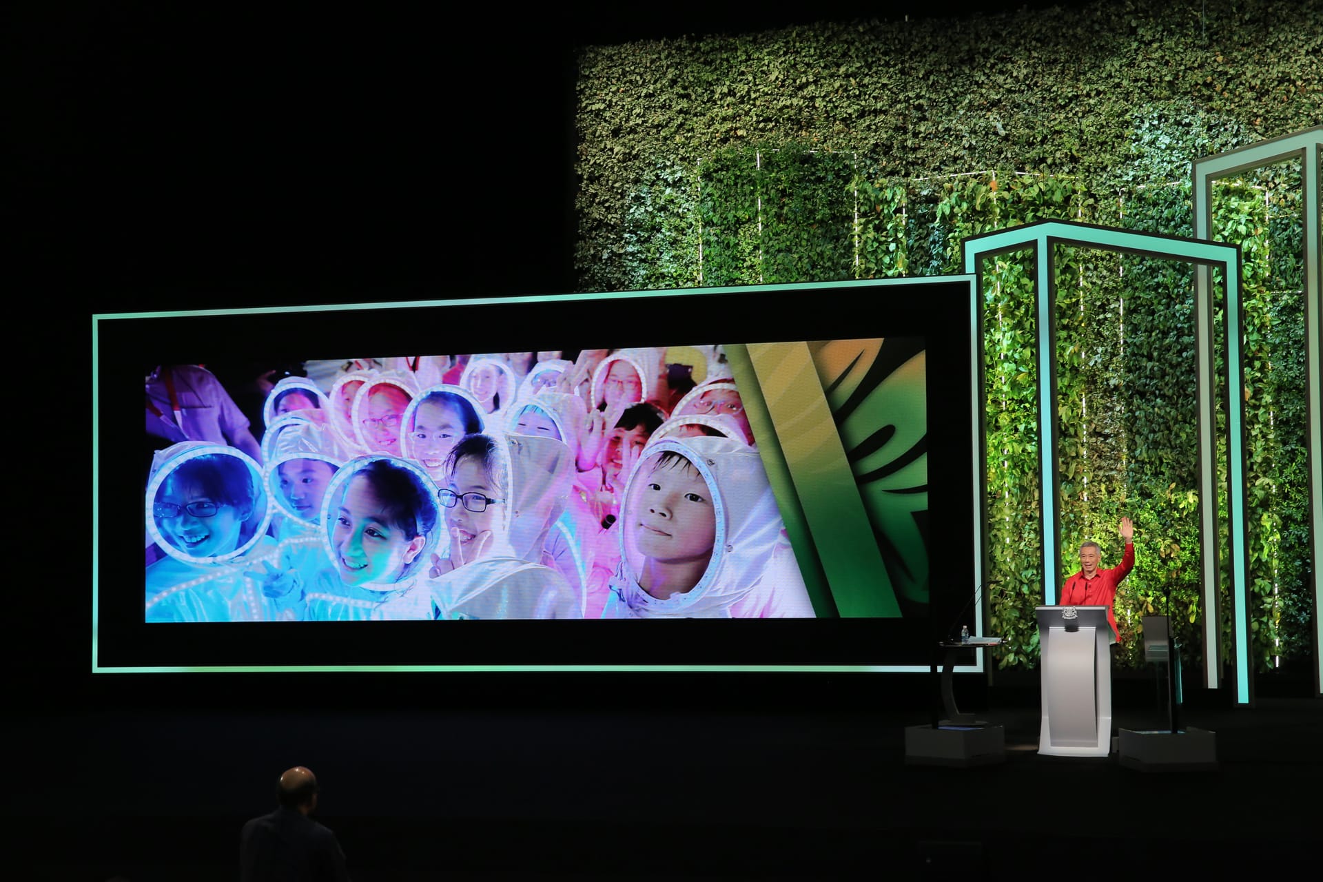 Speaker waves by screen showing kids in white outfits, backdrop of green wall and square frames.